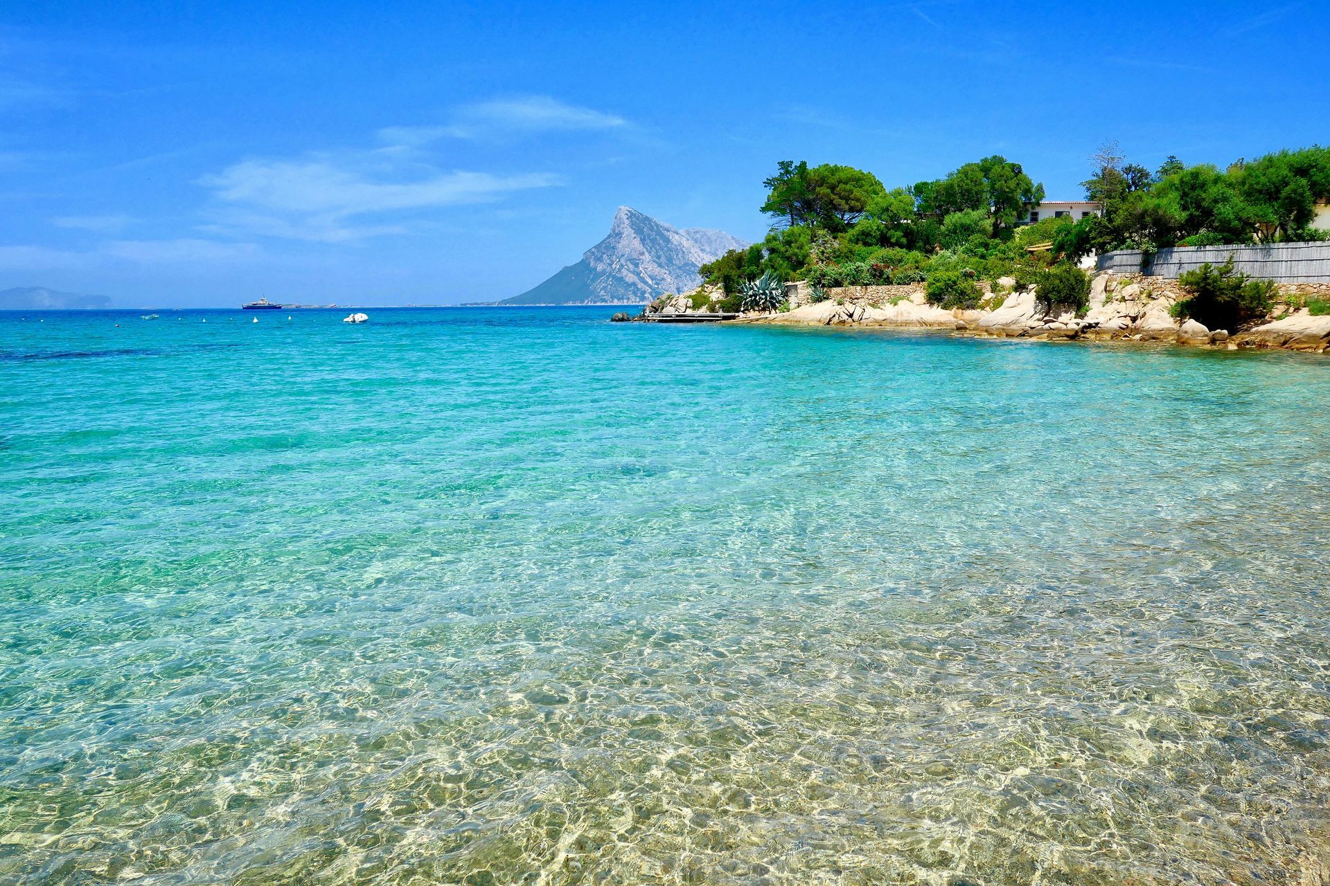 A beach with a boat in the water and a mountain in the background in Sardinia, Italy.