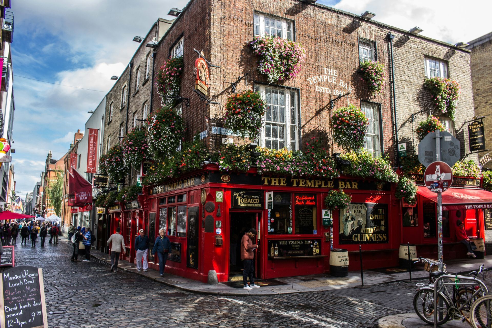 Red Temple Bar pub in Dublin, Ireland, on a cobblestone street, with hanging flower baskets, people, and a bike.