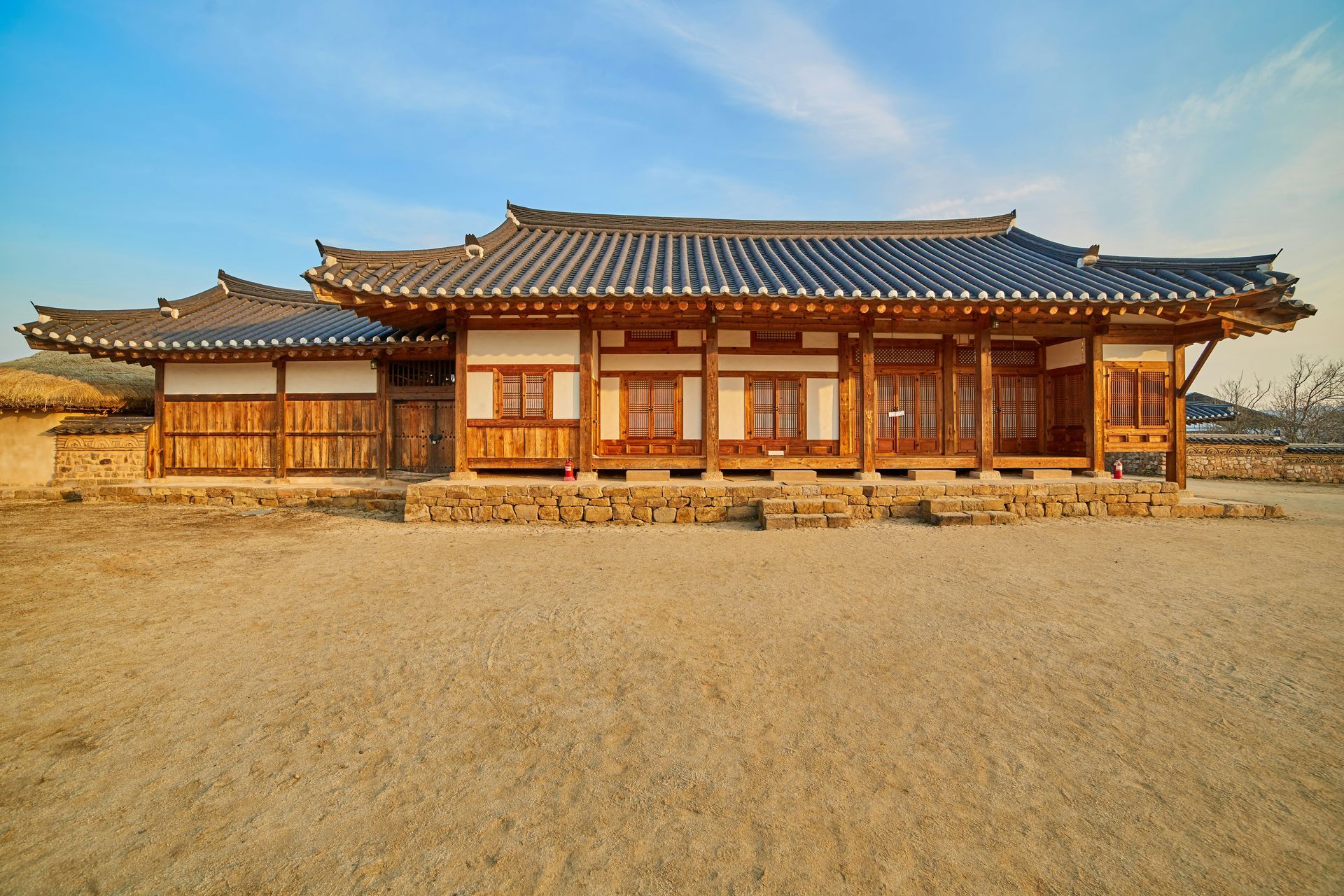 Traditional Korean hanok house with tiled roof and wooden framework on a sandy ground, under a blue sky.