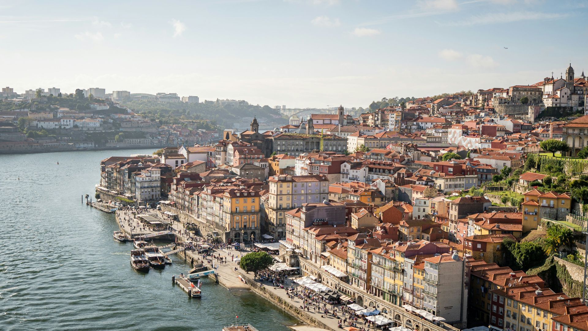 Cityscape of Porto, Portugal, with red-roofed buildings along a river, boats docked, and a sunny sky.