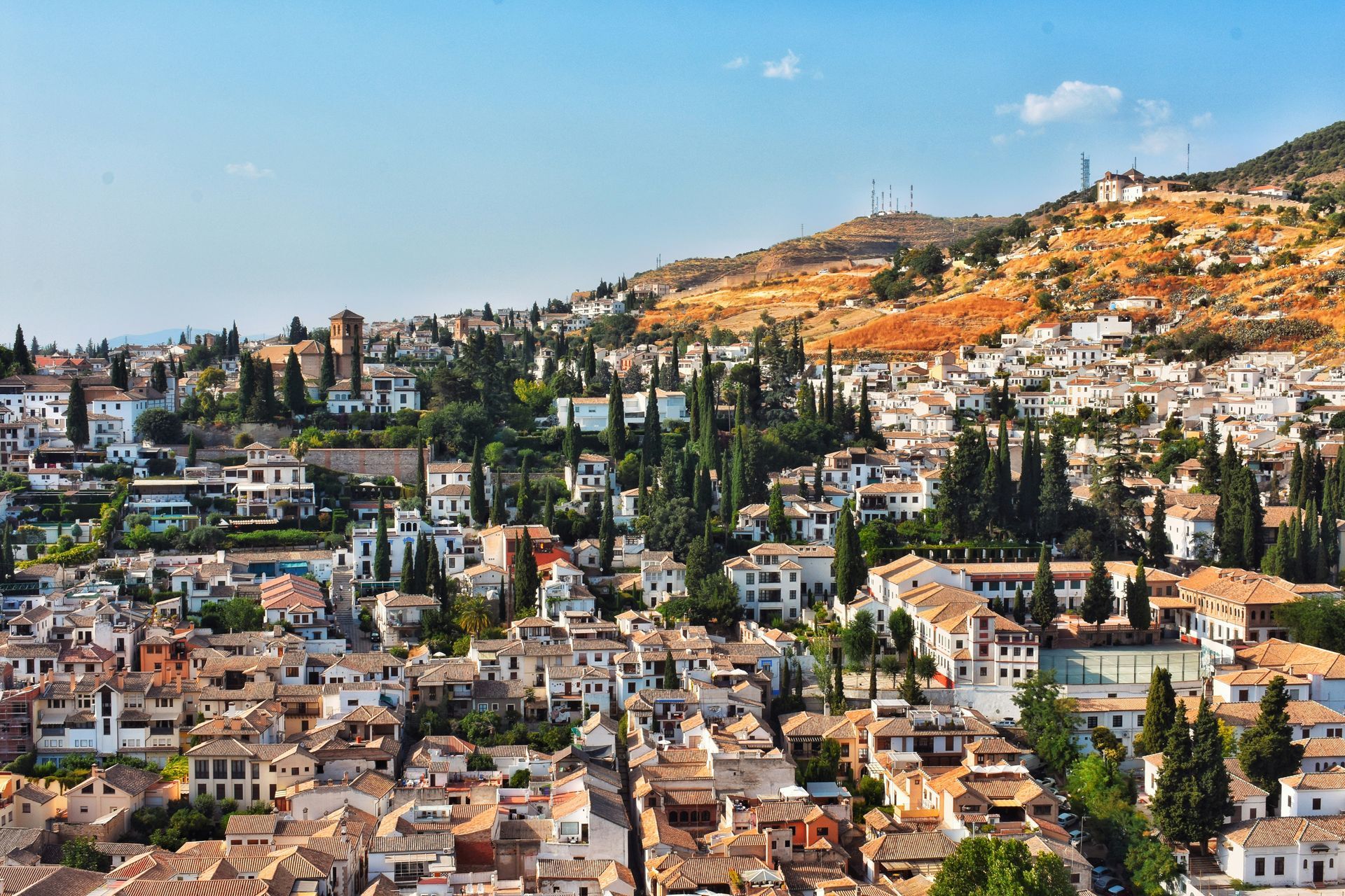 Cityscape of Granada, Spain, with white buildings, terracotta roofs, and green trees on a hillside under a blue sky.