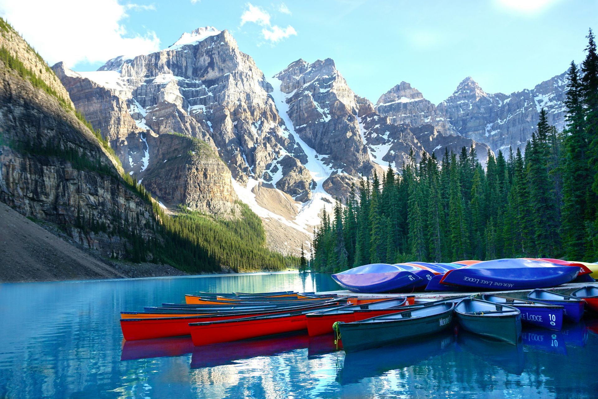 Boats docked on a turquoise lake with snow-capped mountains in the background; a sunny day in a forest in Banff, Canada.