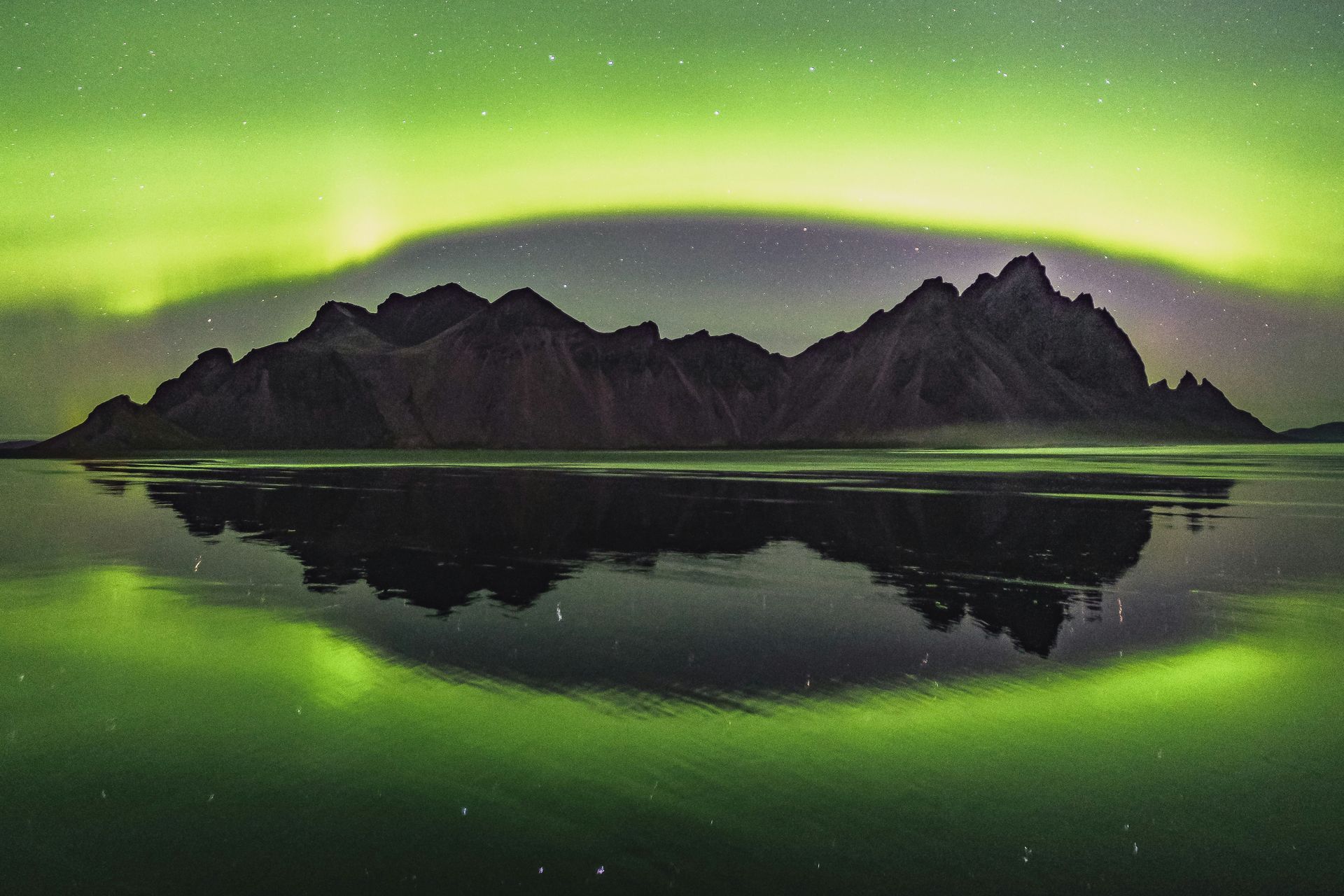 Green aurora borealis over dark mountains reflected in water in Stokksnes, Iceland.