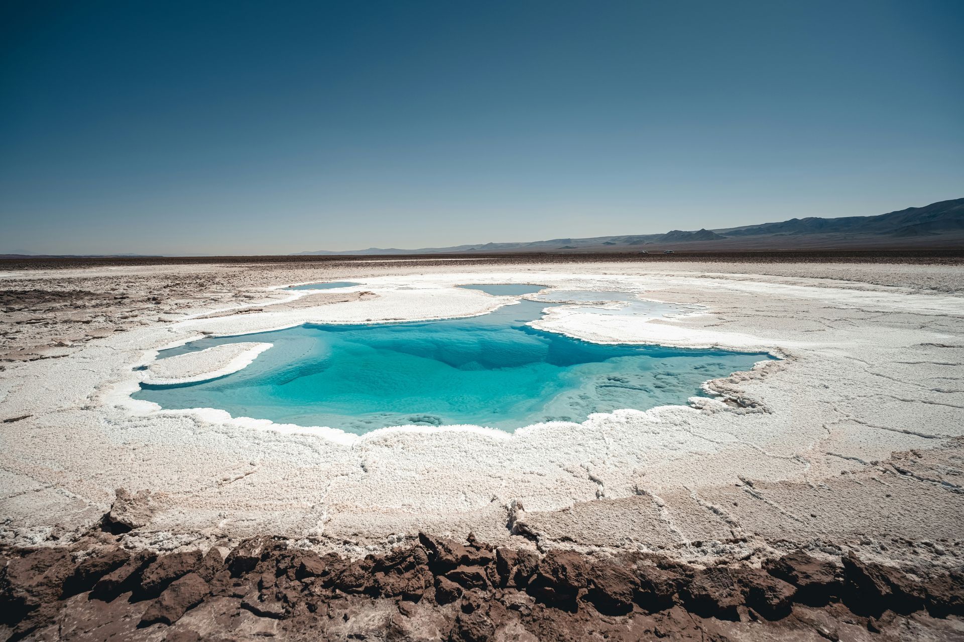 Bright turquoise water in a salt flat, surrounded by white salt and arid landscape, under a blue sky in the Atacama Desert, Chile.