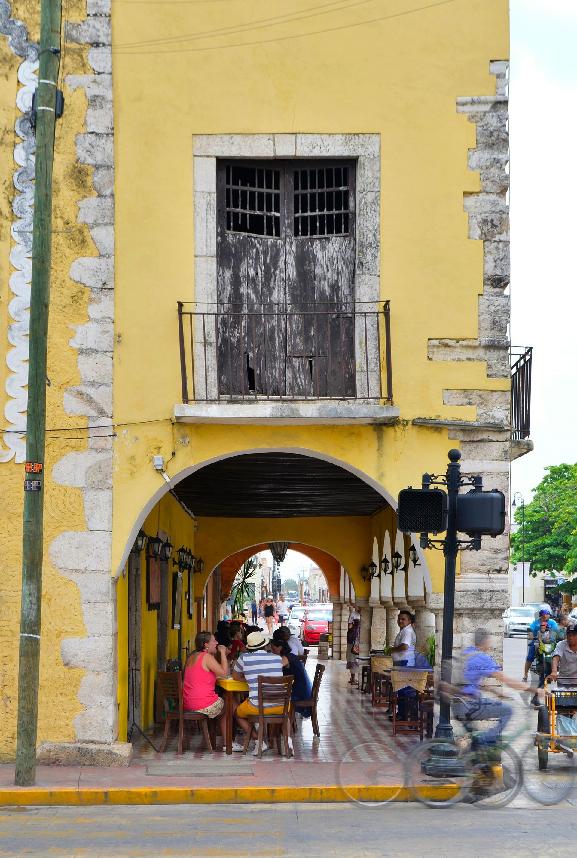 Yellow building with an arched walkway. People sit at outdoor tables on the street in front in Valladolid, Mexico.
