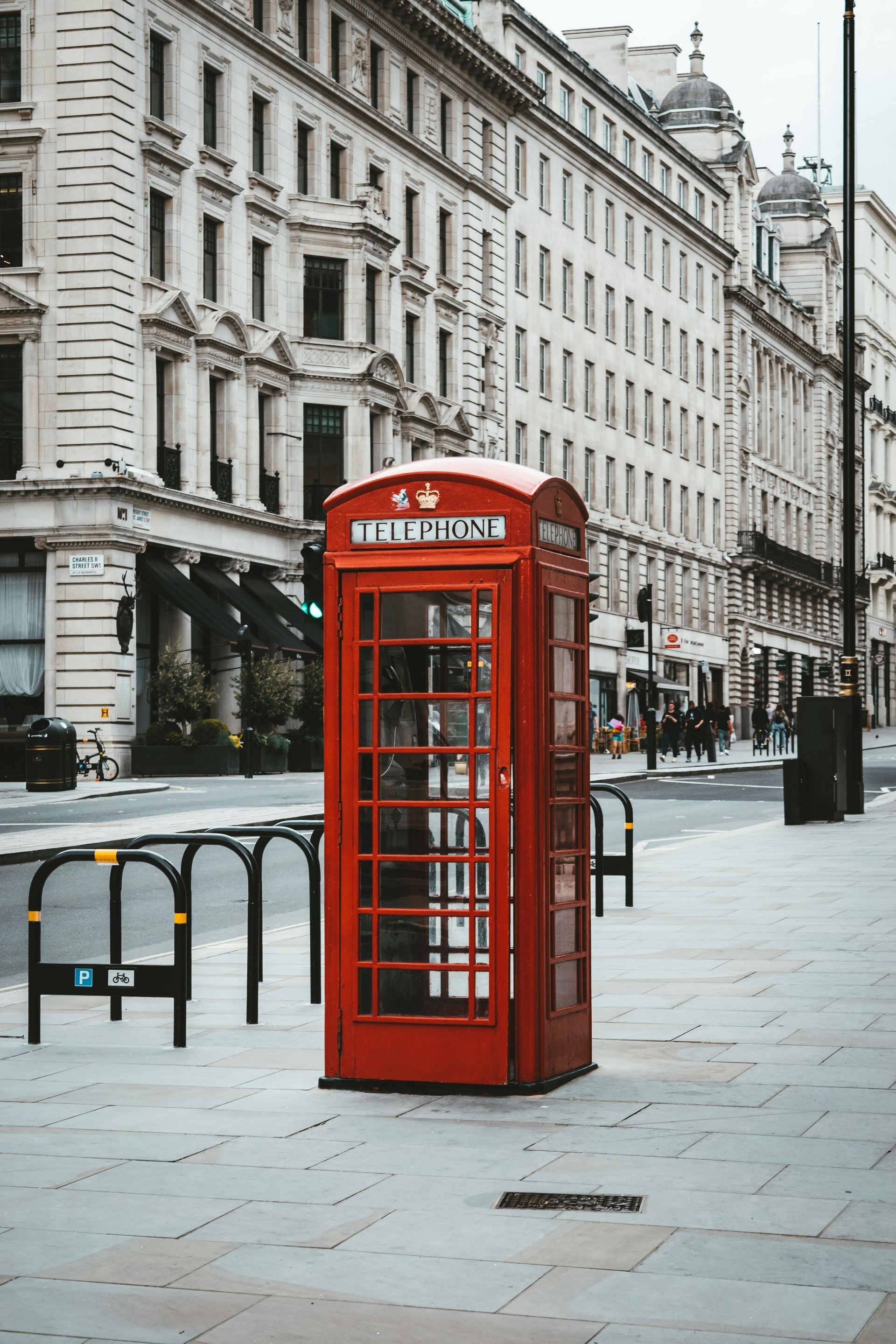 Red telephone box on a London sidewalk, surrounded by stone buildings and traffic barriers.