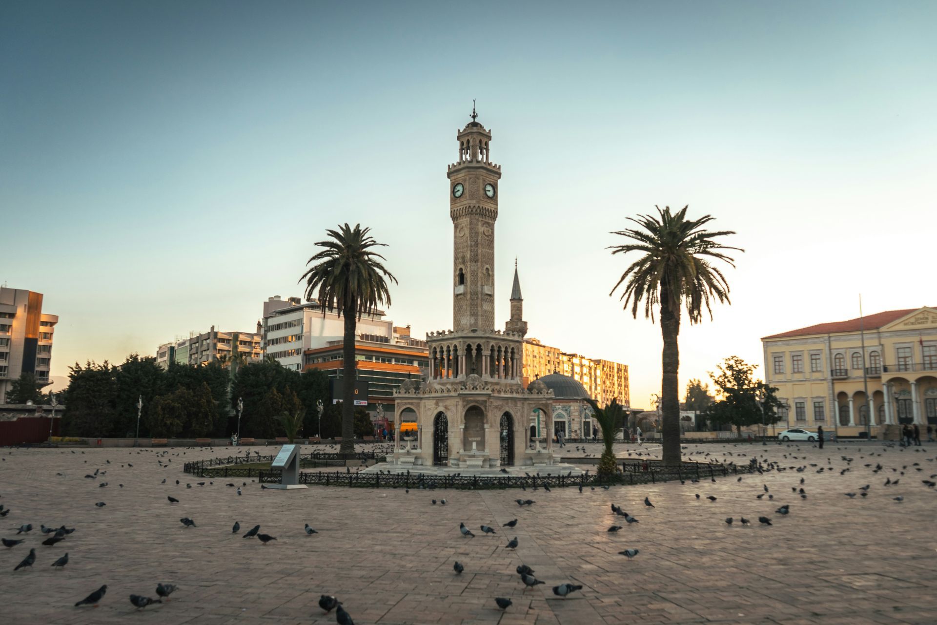 Clock tower in Konak Square, Izmir, Turkey, with palm trees and pigeons at sunset.