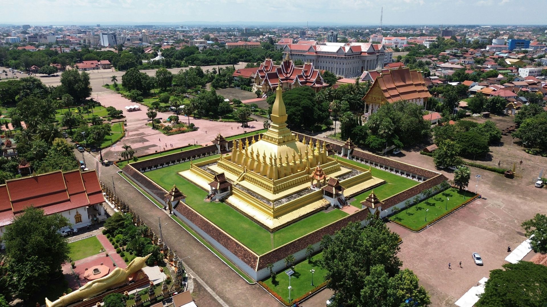 Golden That Luang stupa in Vientiane, Laos. Surrounded by a wall and green lawns. Cityscape in the background.