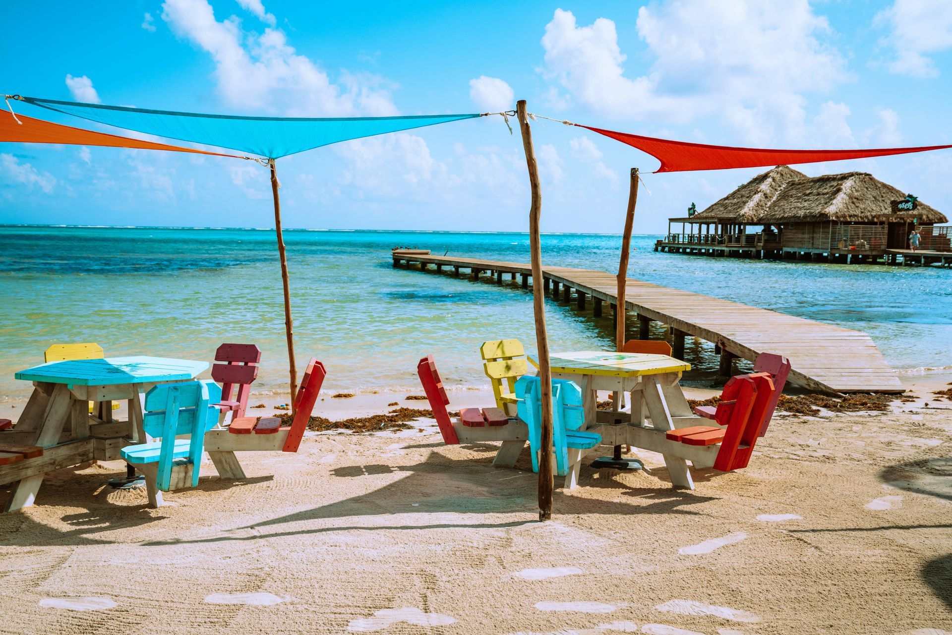 Colorful picnic tables with shade sails on a sandy beach. A wooden pier leads to a building over the water in Ambergris Caye, Belize.
