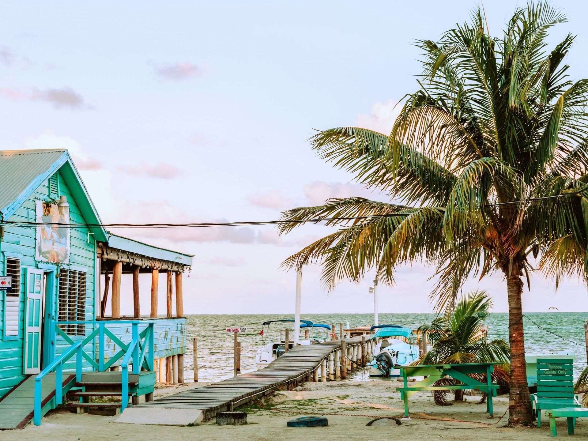 A turquoise beachside building with a wooden pier, palm trees, and ocean view under a pastel sky in Belize.