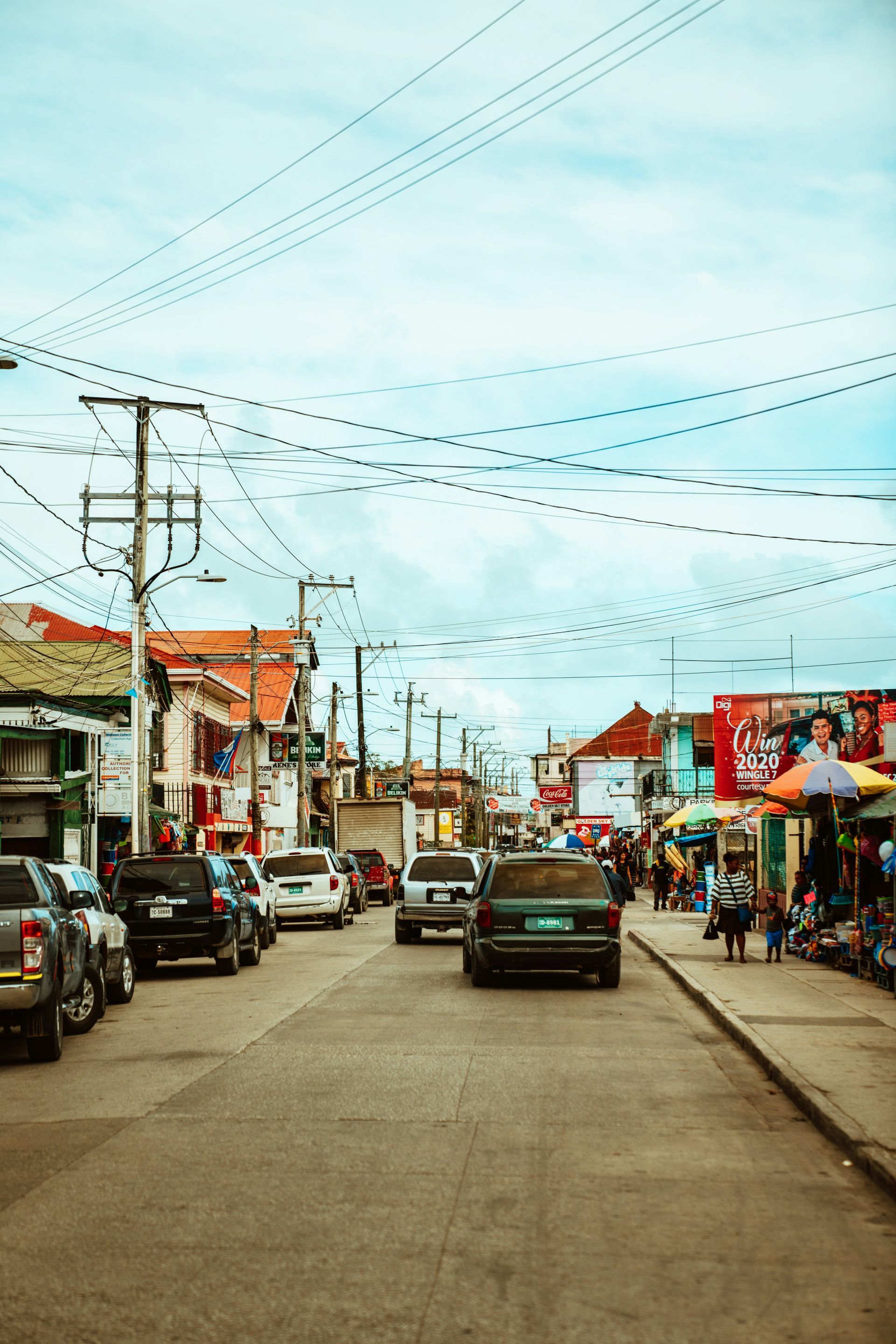 Street scene in a bustling town with cars and buildings, overhead power lines, and people walking along the sidewalks in Belize City.