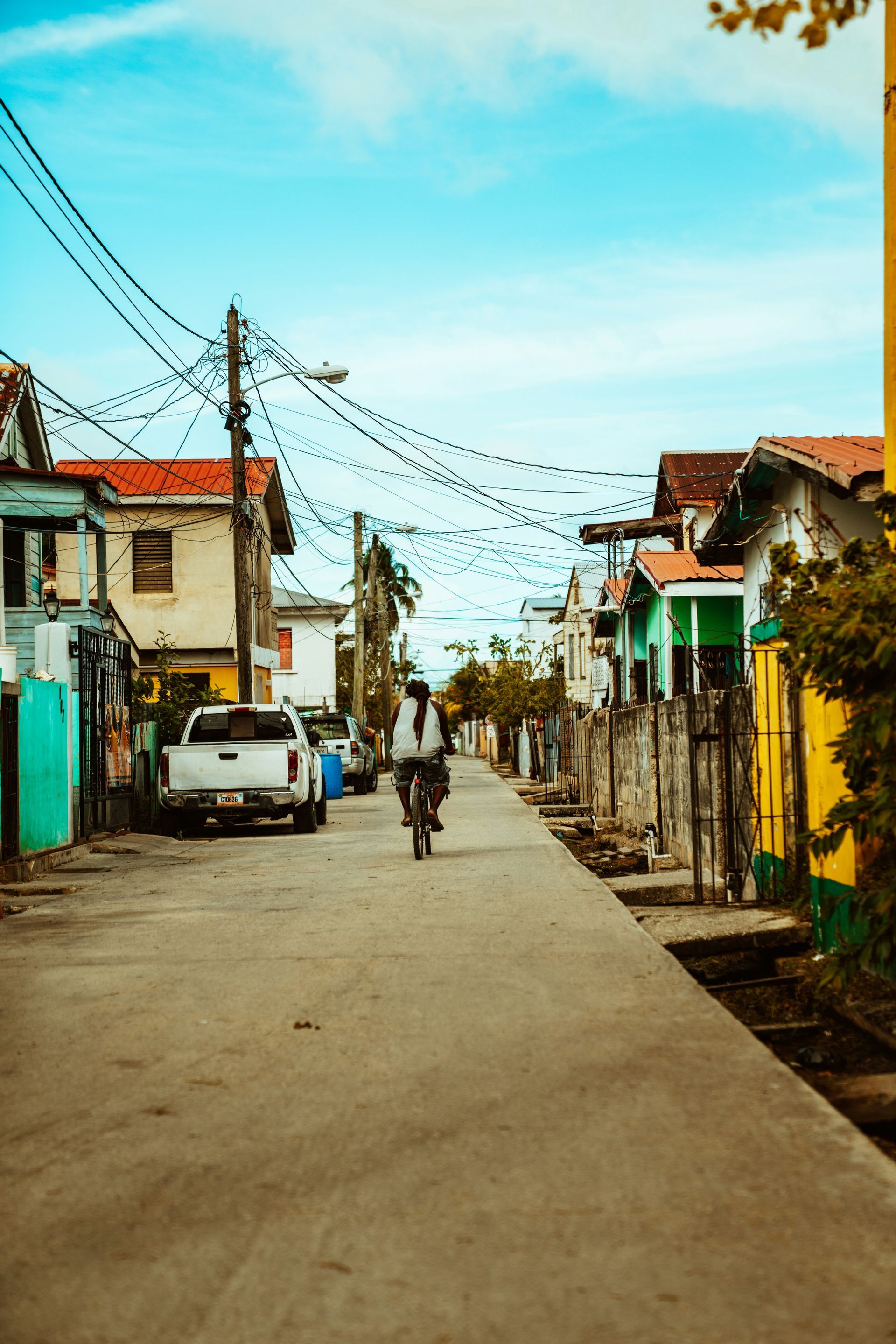 A cyclist rides down a narrow street lined with colorful houses under a bright blue sky in Belize City.