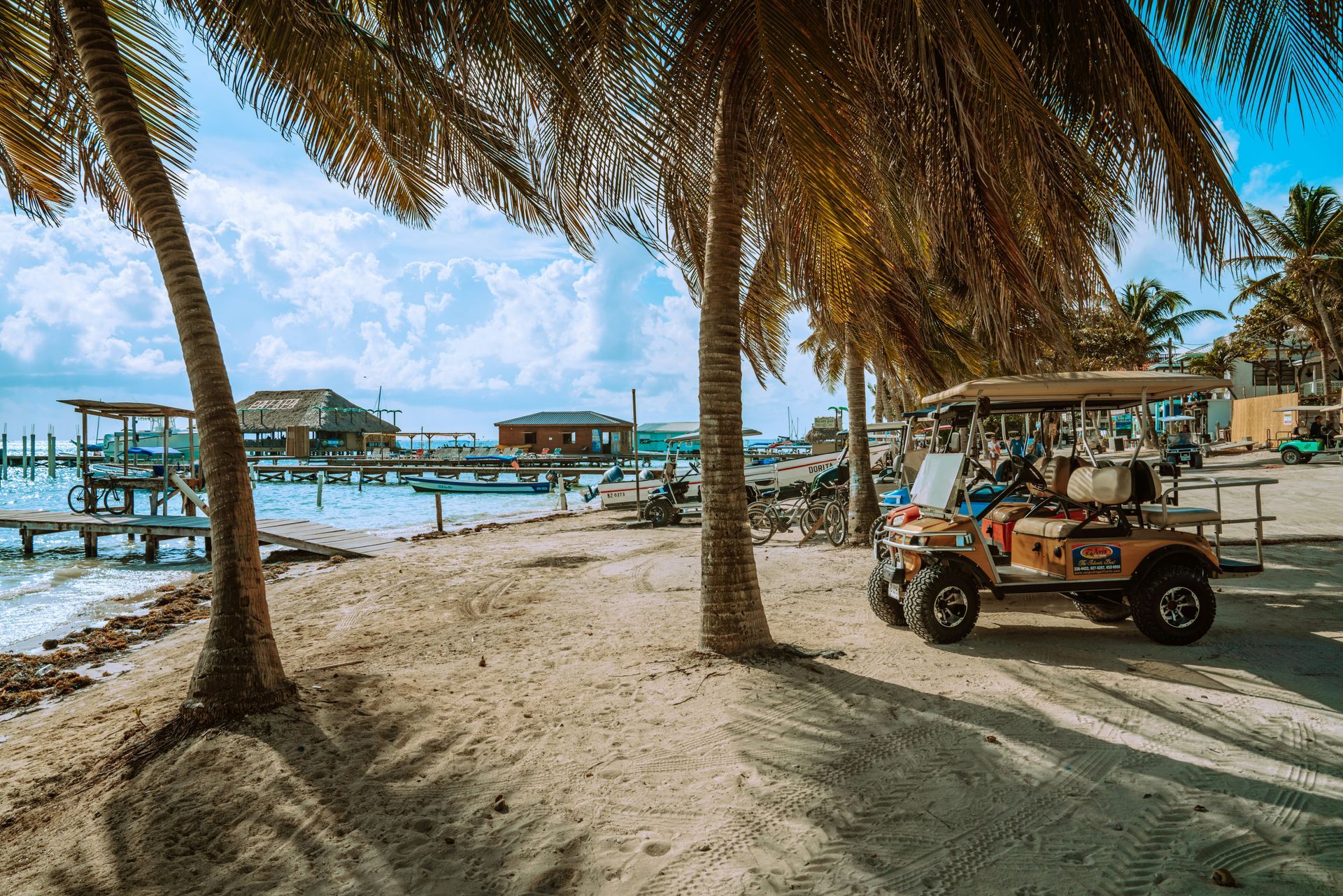 Beach scene with palm trees, a golf cart, and buildings on stilts over the water. Sunny day in Ambergris Caye, Belize.