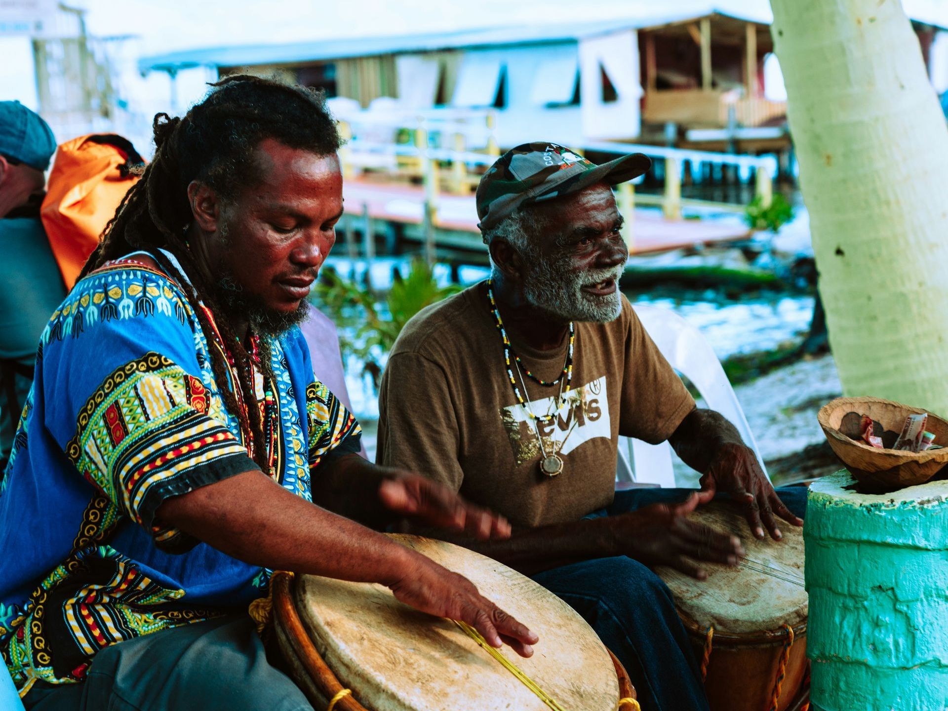 Two men playing drums outdoors near a waterfront. One wears blue patterned clothing, the other a brown t-shirt in Belize.