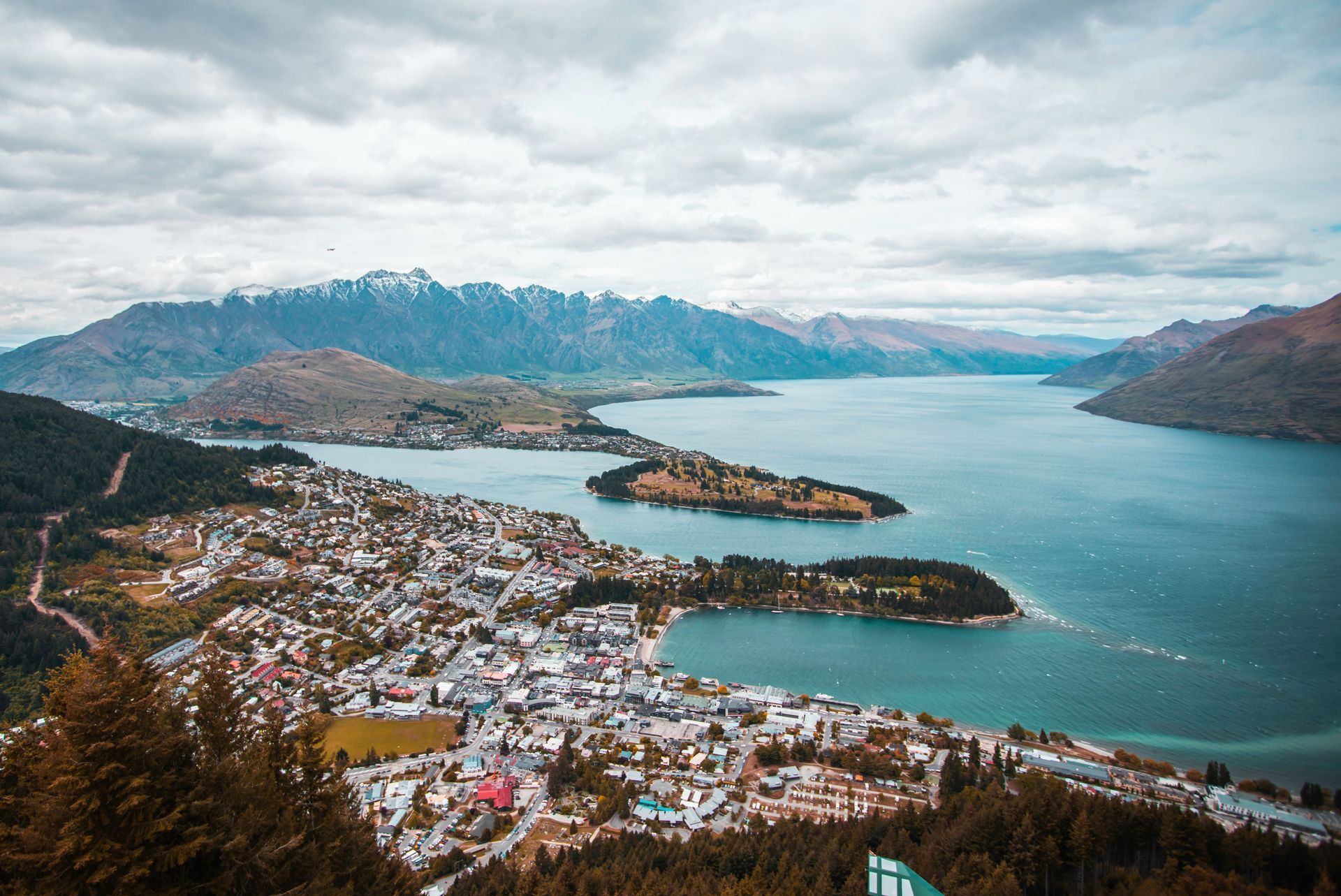 An aerial view of a lake with mountains in the background and Queenstown, New Zealand in the foreground.