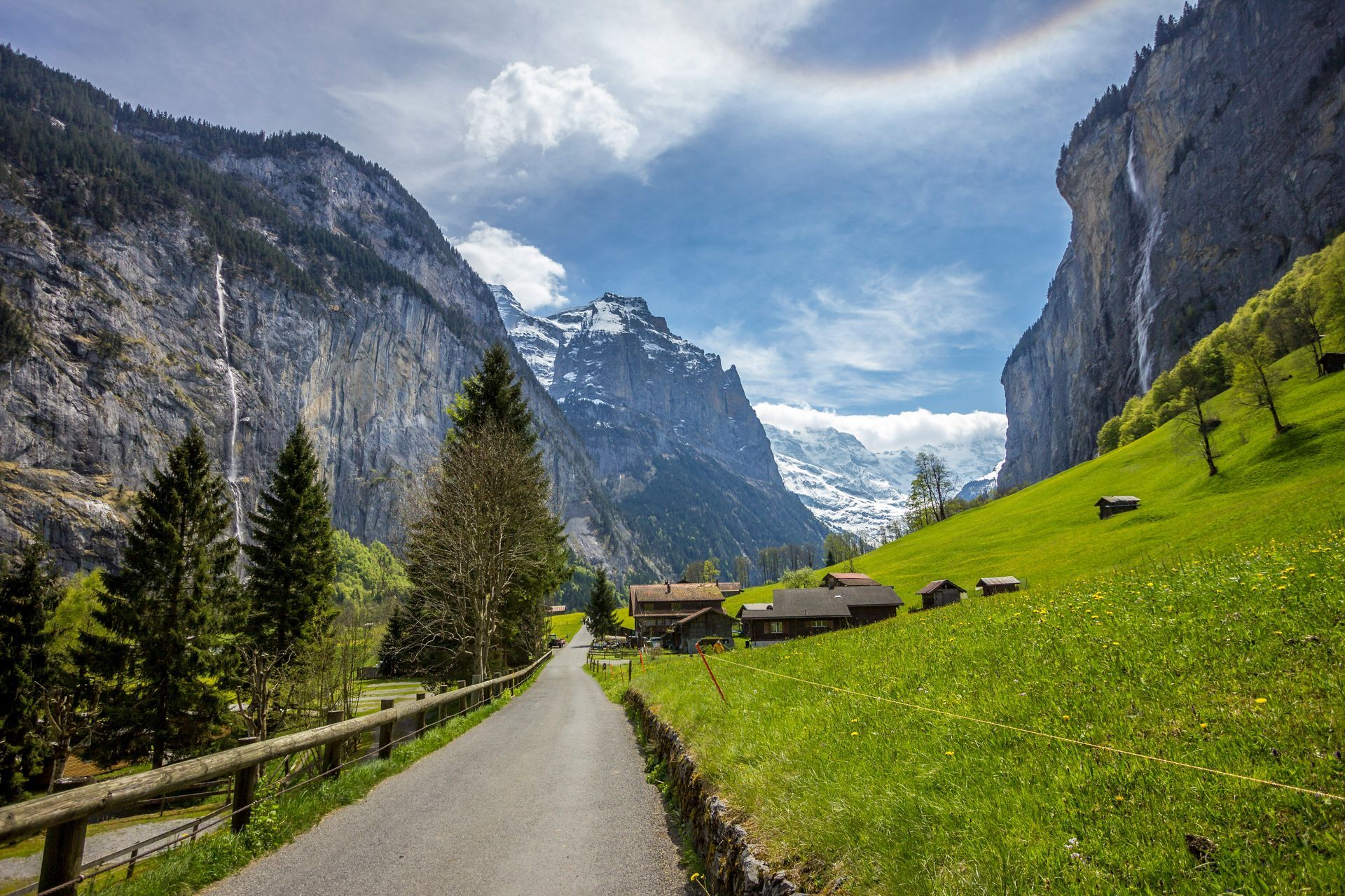Road leading to a village nestled in a valley between mountains and waterfalls, under a partly cloudy sky in Lauterbrunnen.