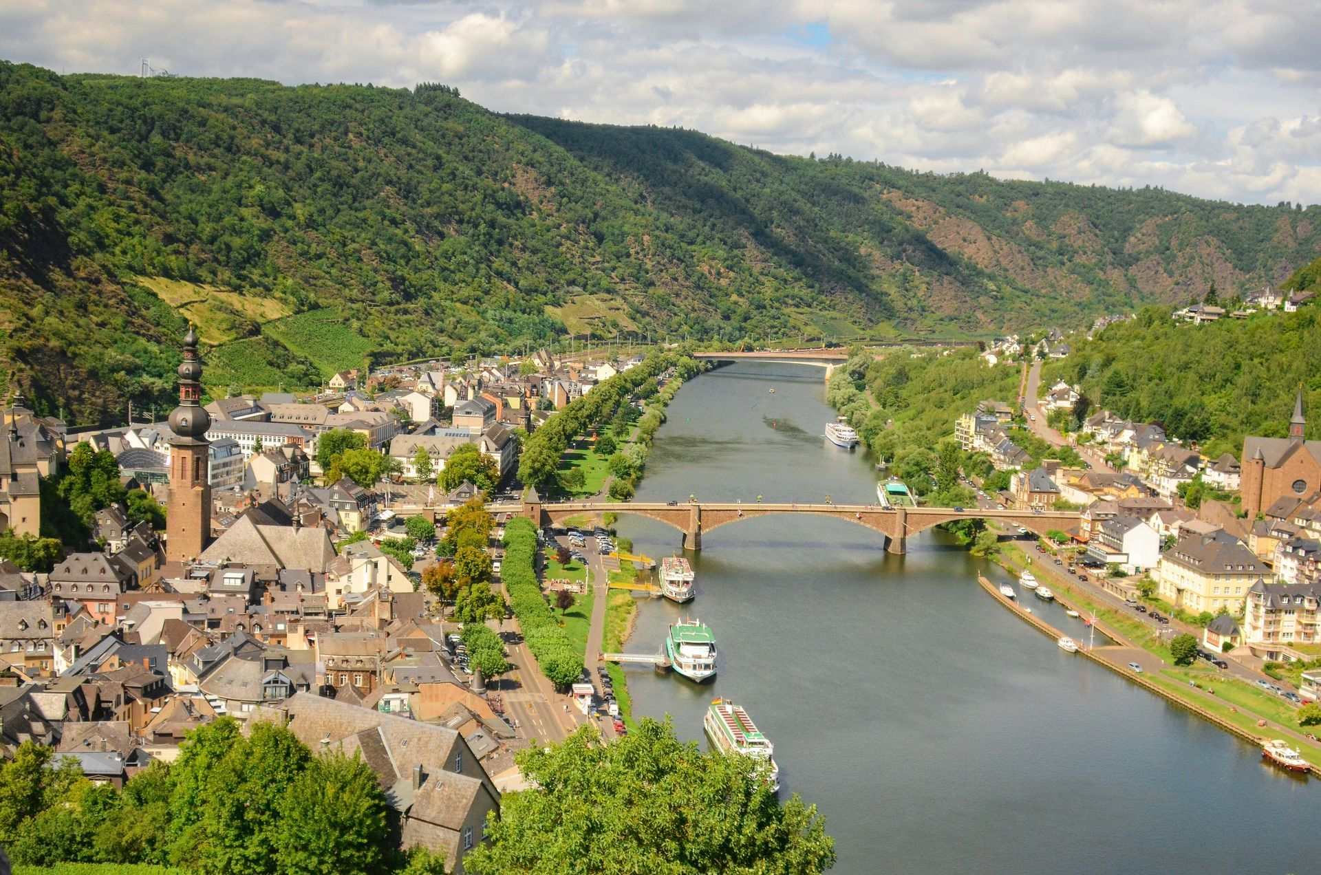 Town of Cochem, Germany along a river with a bridge, surrounded by green hills. Boats are docked on the river.