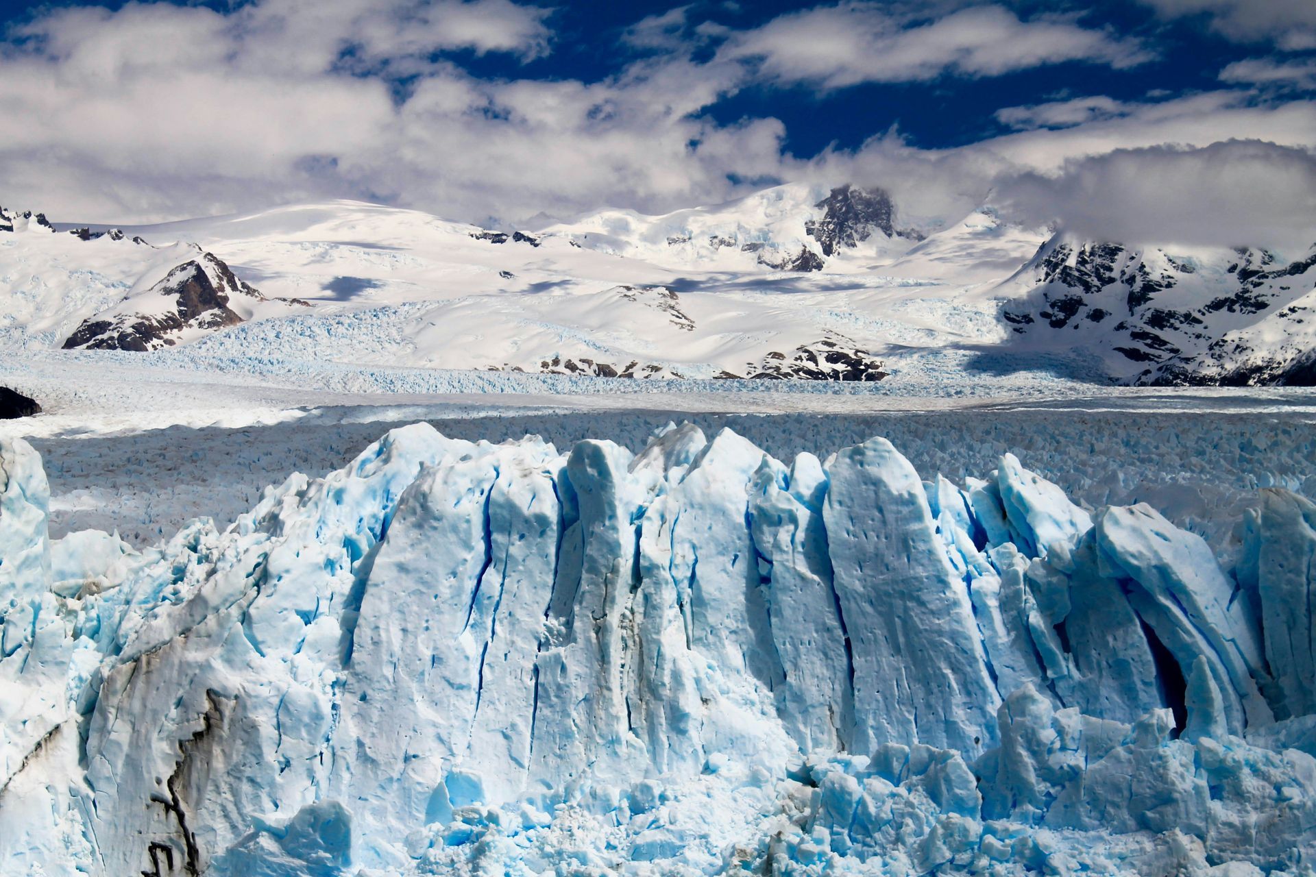 Glacier with jagged blue ice in front of snow-capped mountains at Parque Nacional Los Glaciares in El Calafate, Argentina.