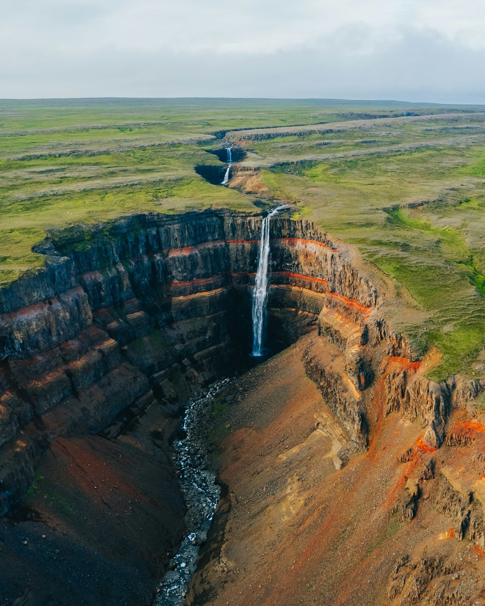 The Hengifoss Waterfall cascades down a layered rock cliff into a narrow canyon, surrounded by green fields in Iceland.