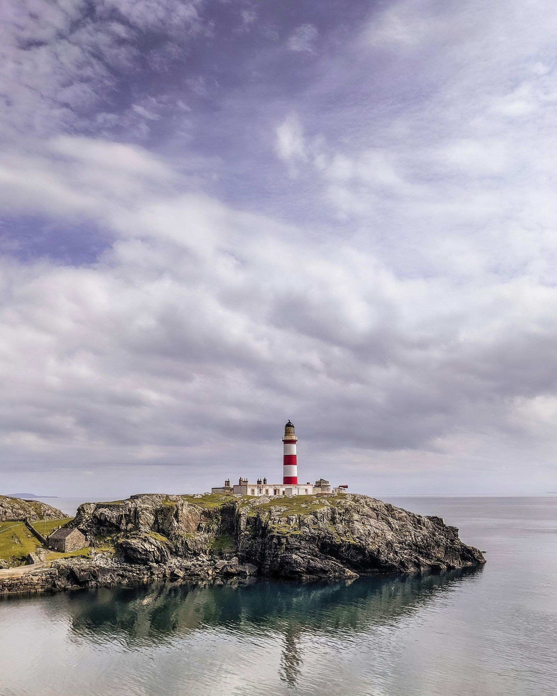 Lighthouse with red and white stripes sits atop a rocky island, reflected in calm water on the Hebrides Islands, Scotland.