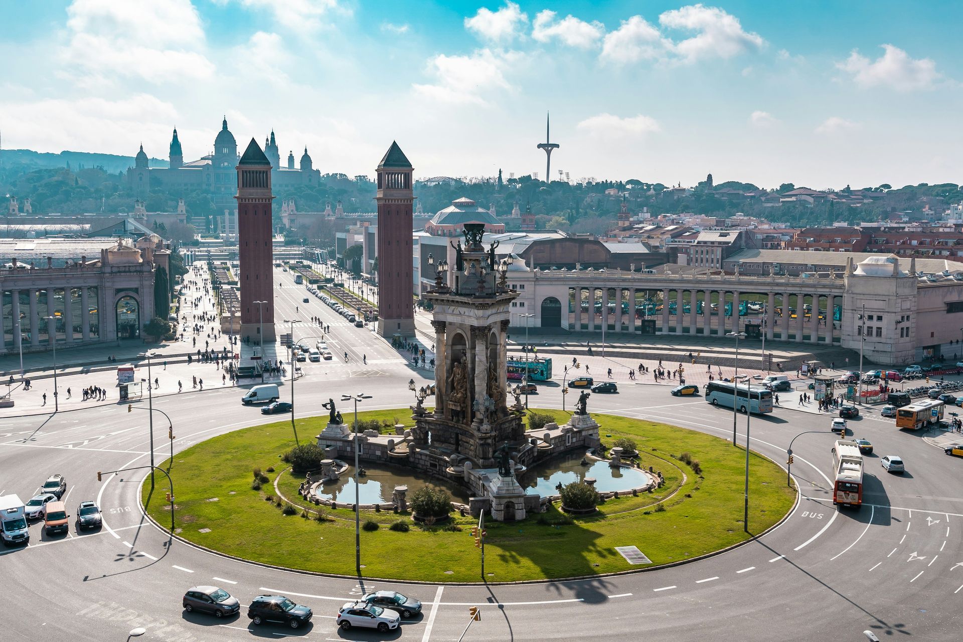 A wide shot of Plaça Espanya in Barcelona, Spain. Features a roundabout with a fountain, tall brick towers, and buildings.