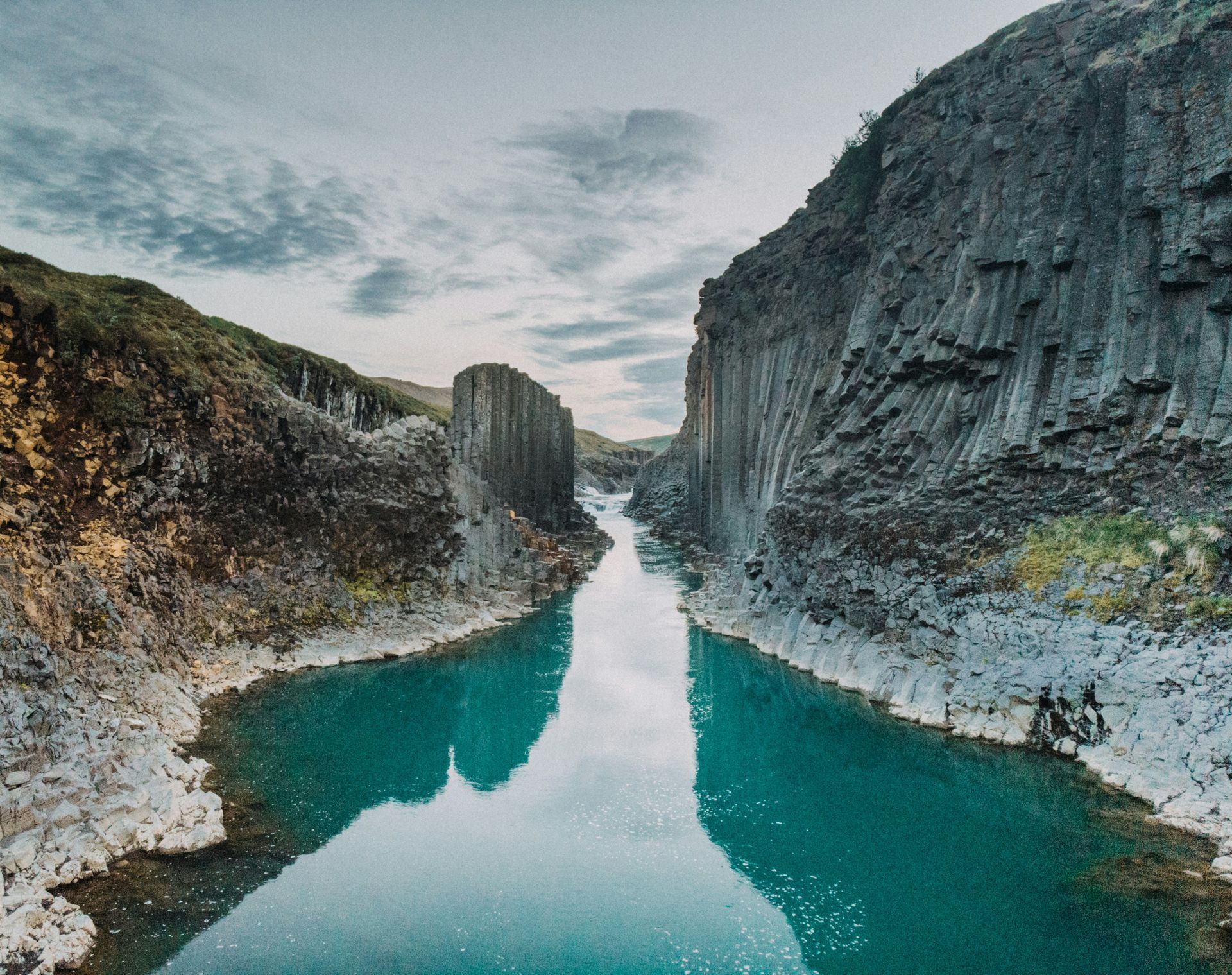 A turquoise river cuts through tall, dark rock formations under a cloudy sky at Stuðlagil Canyon, Iceland.