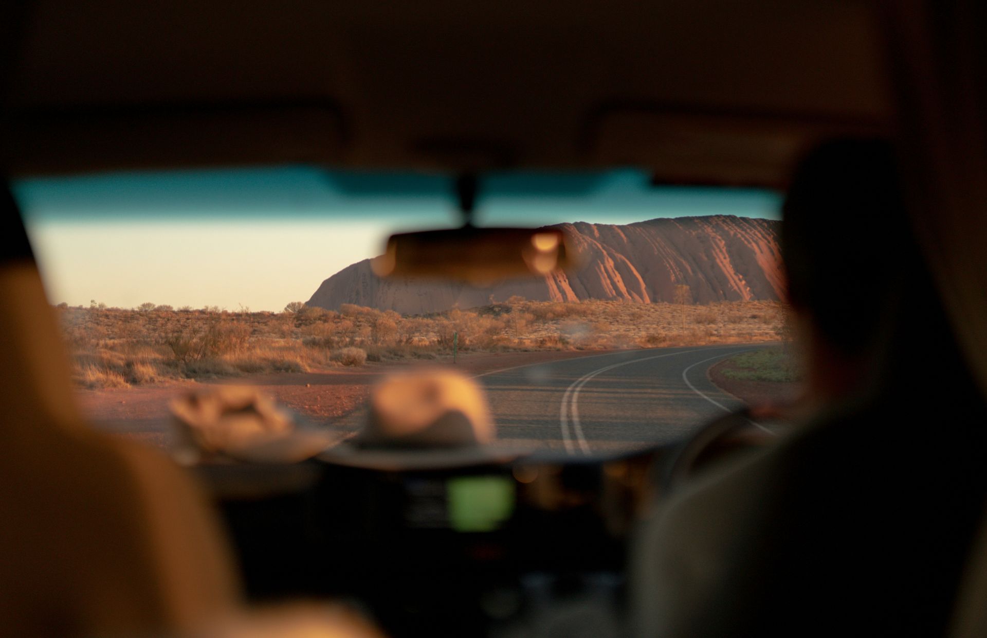 A person is driving a car down a desert road with a view of Uluru Rock in the windshield in Australia. 