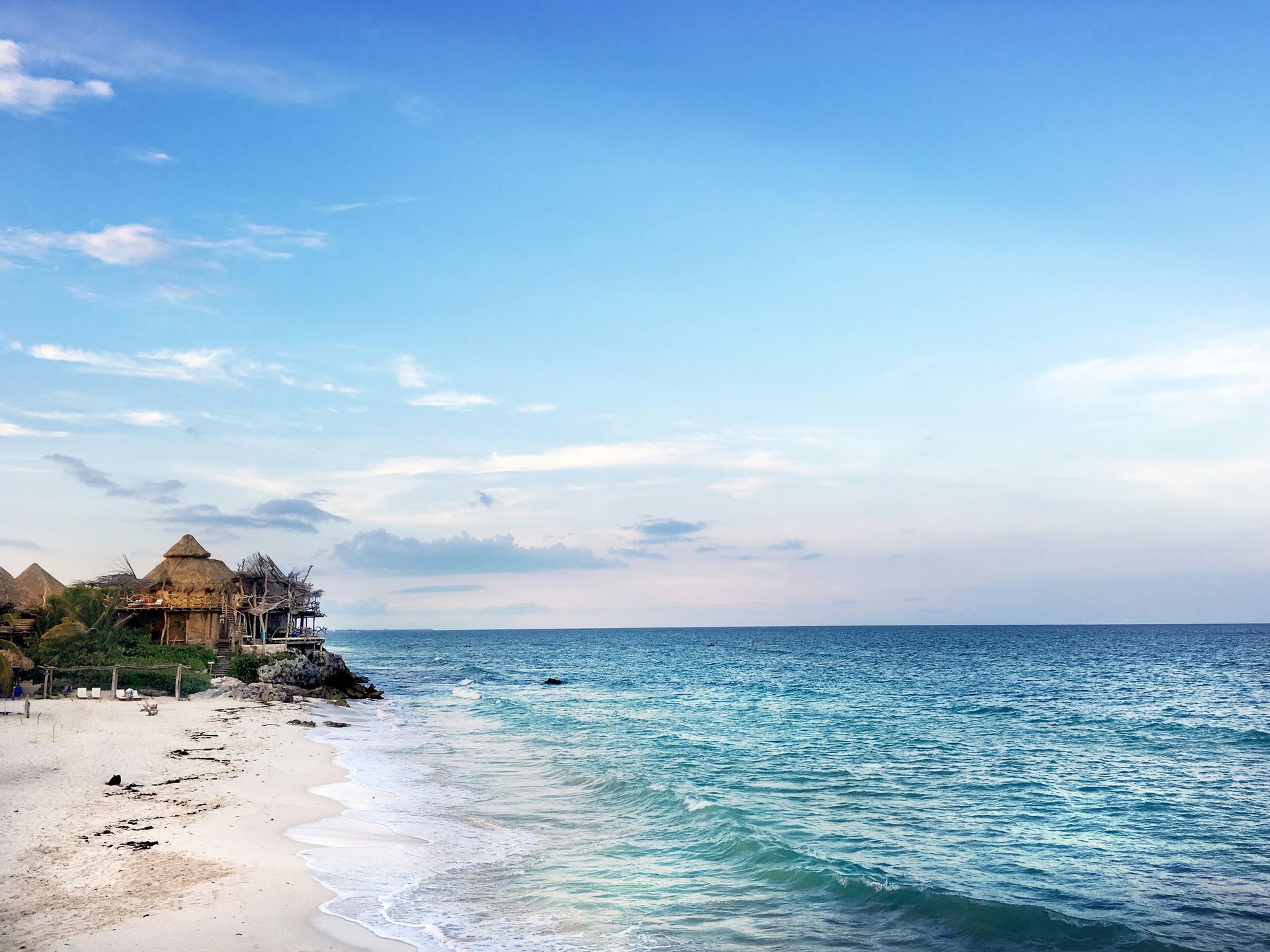 Beach with turquoise water, white sand, and buildings with thatched roofs in Tulum, Mexico.