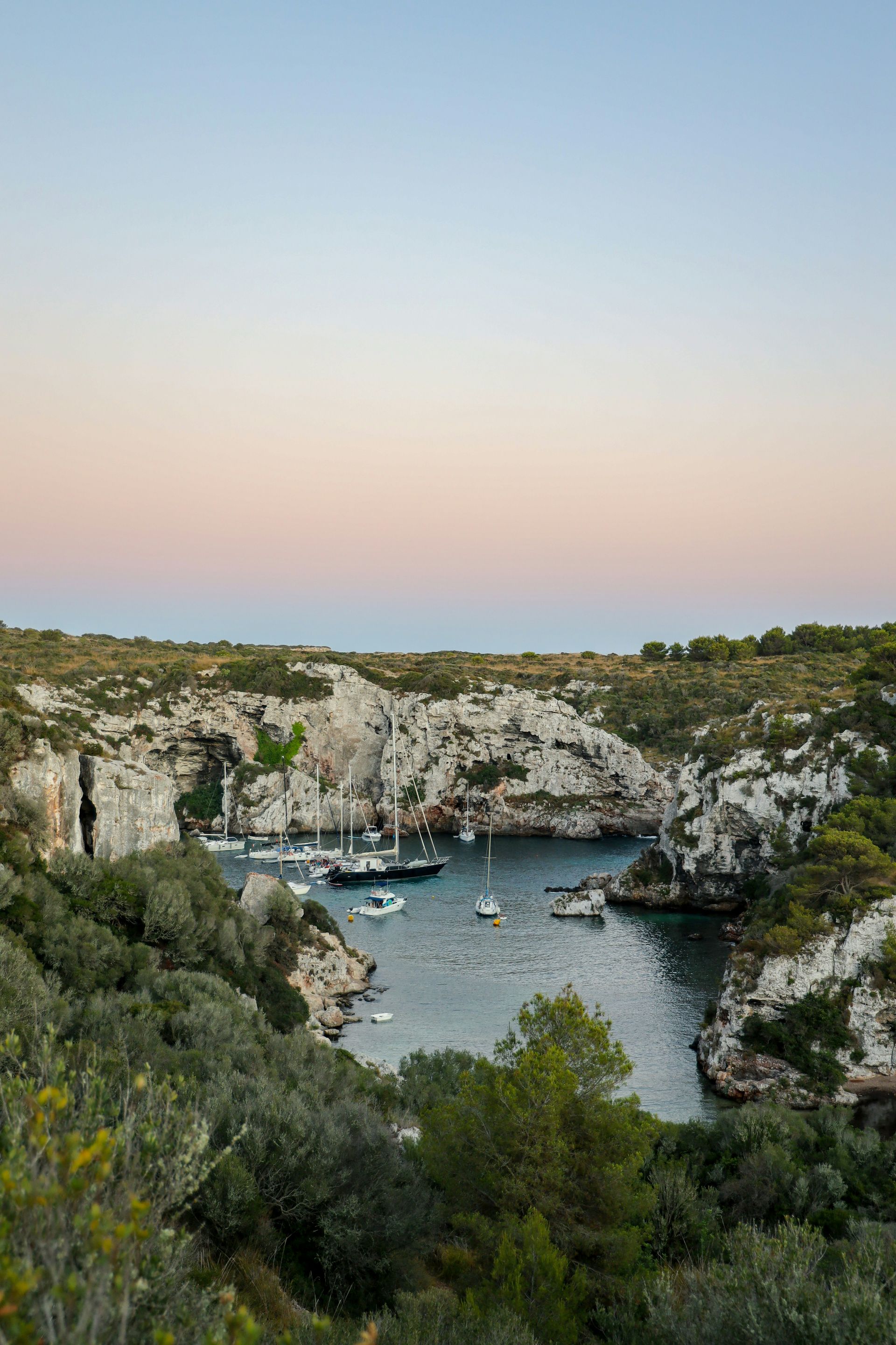 Coastal inlet with boats, surrounded by cliffs and vegetation, under a colorful dusk sky in Menorca, Spain.