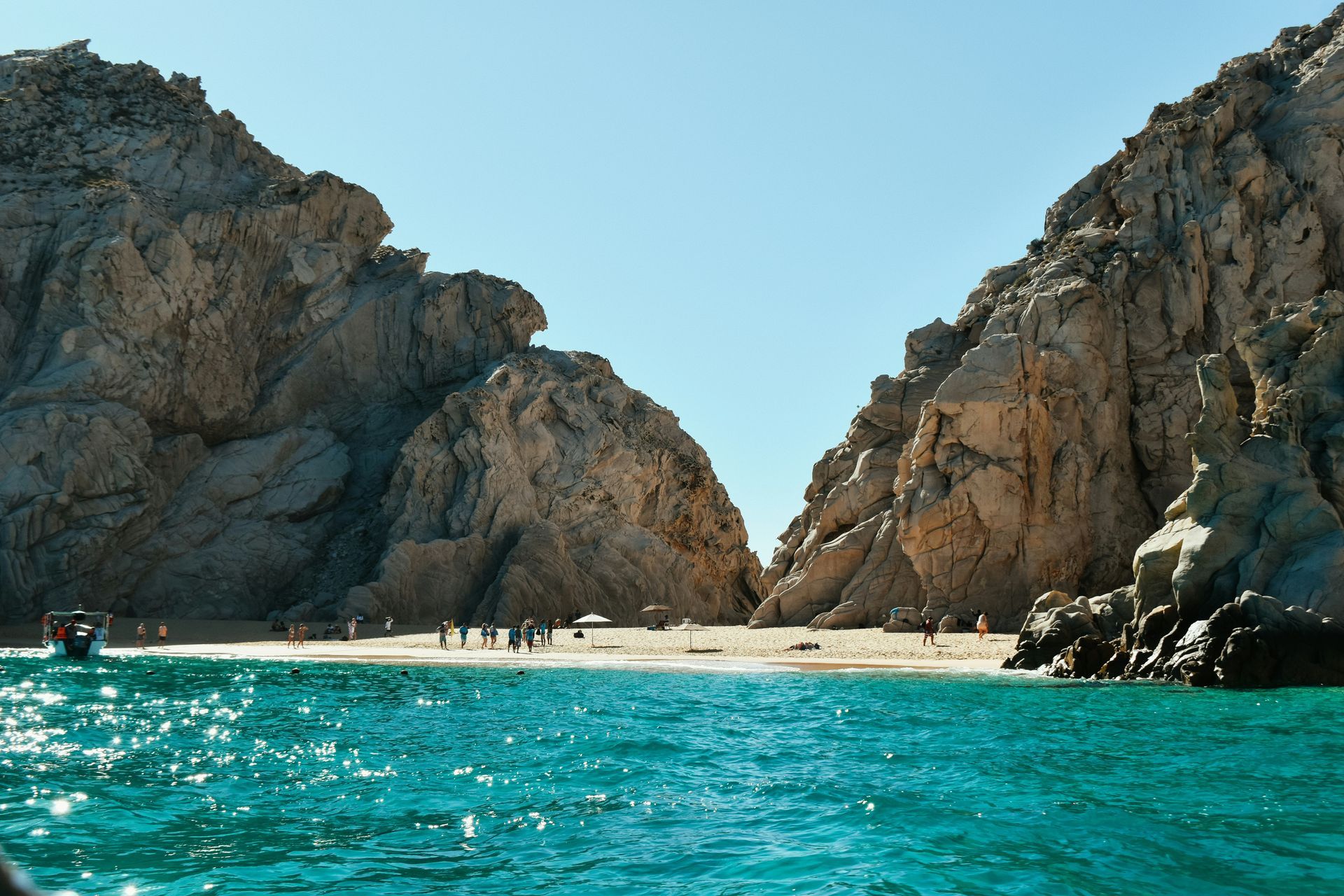 Rocky cliffs frame a small beach with turquoise water, with people visible on the sand in Cabo San Lucas, Mexico.