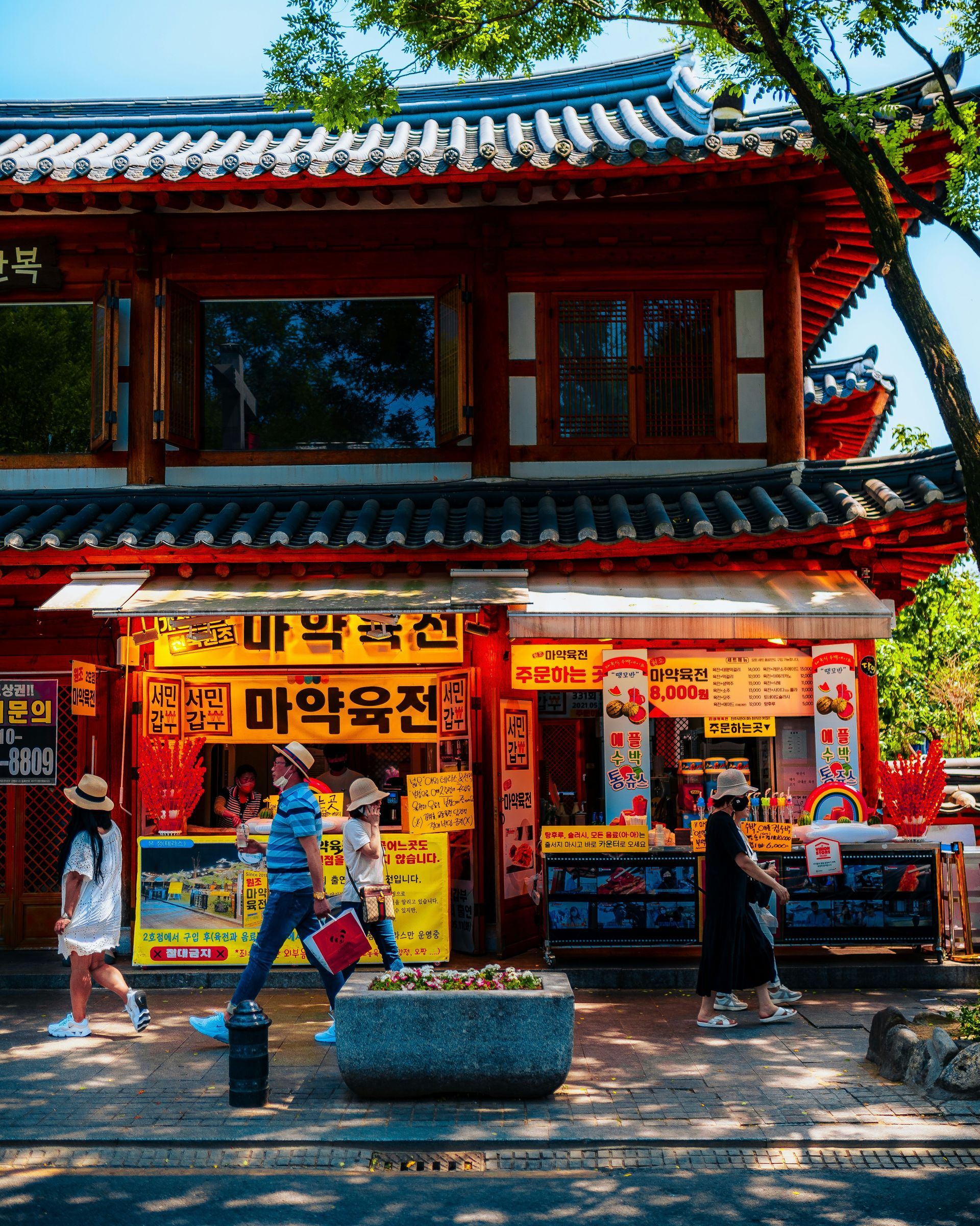 Red-roofed building with food stalls; people walking on a street, some wearing masks in Jeonju Hanok, South Korea.