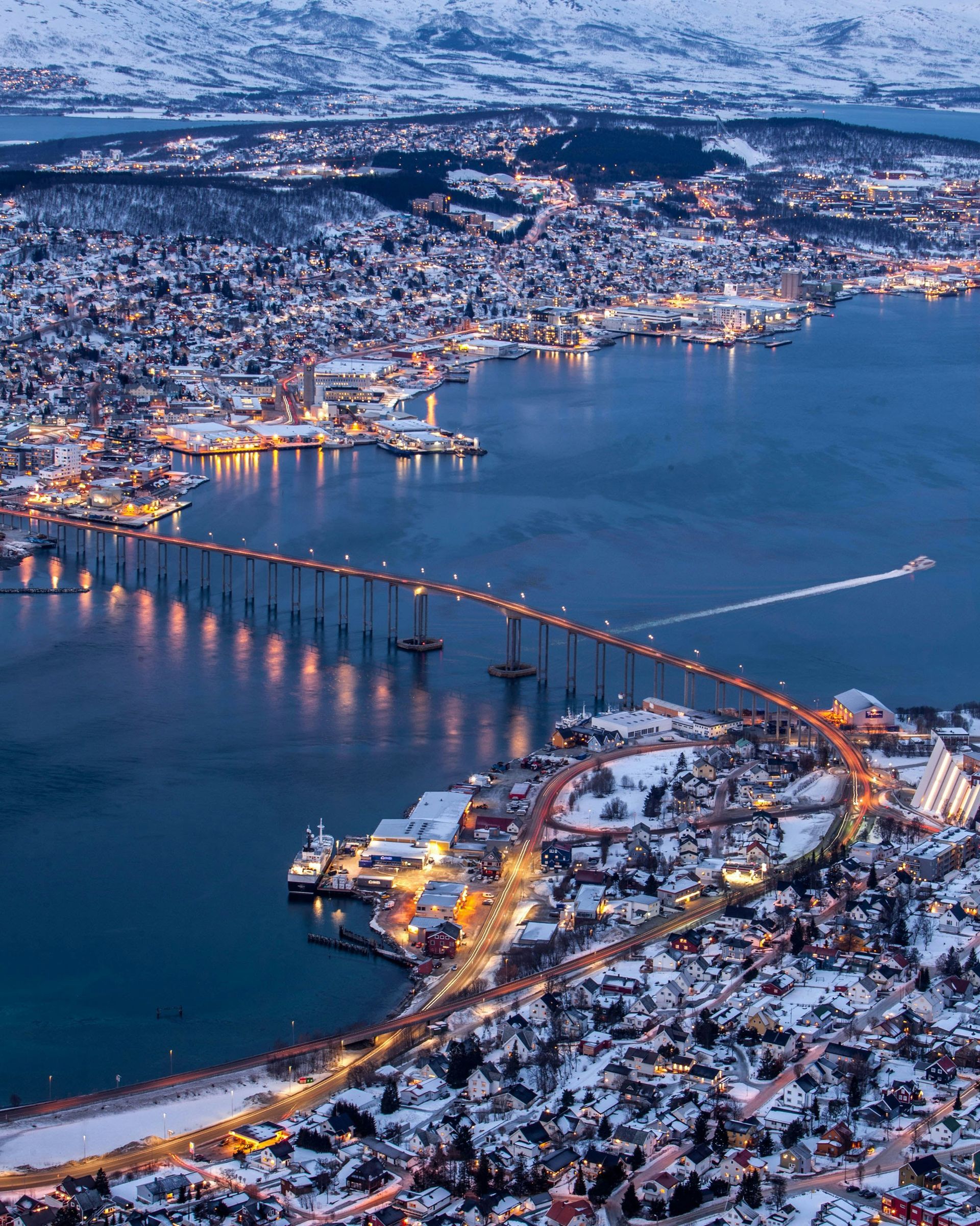 Tromsø, Norway, at dusk, lit bridge over water, snow-covered buildings, mountains in the background.