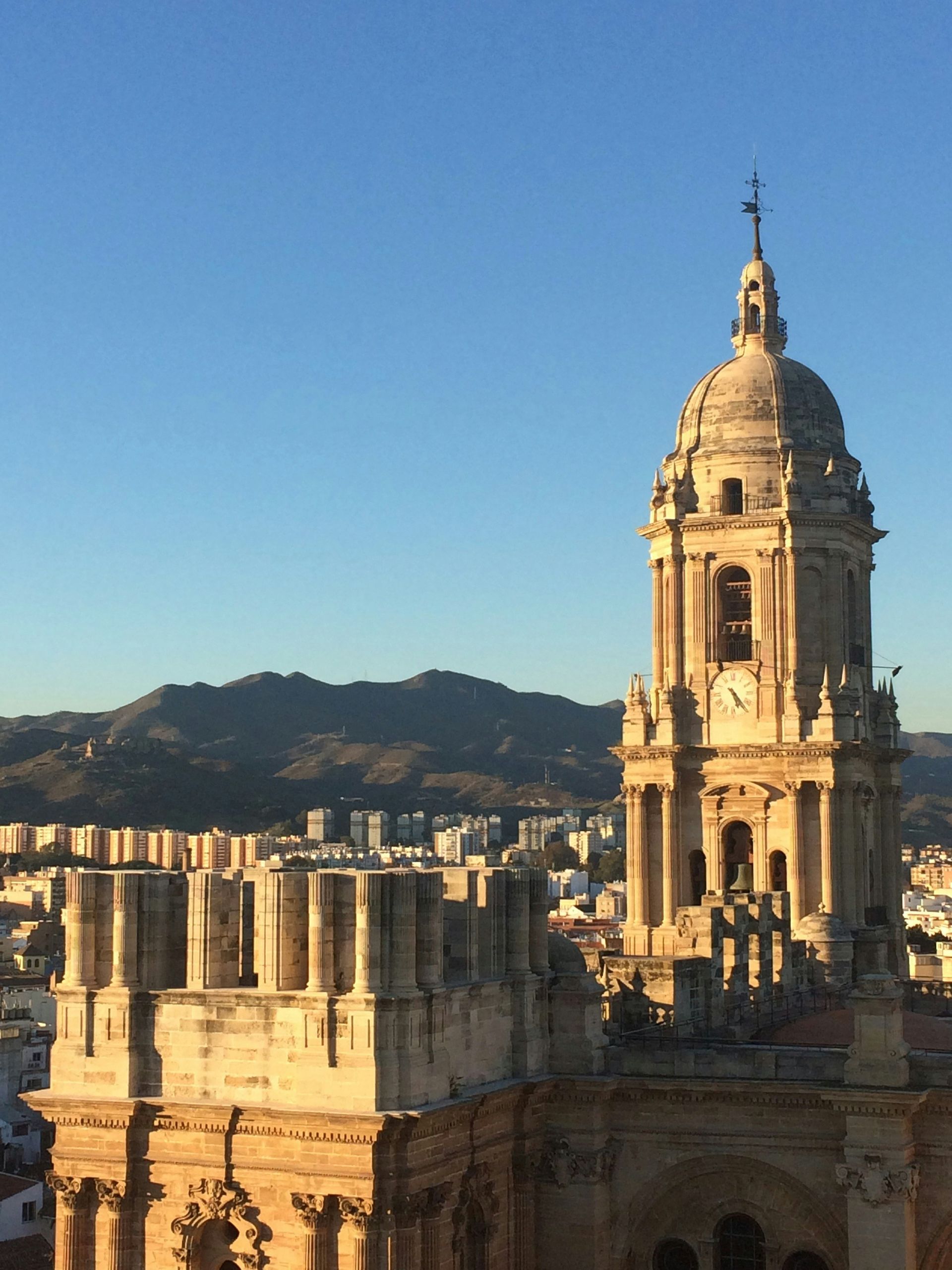 Tower of Malaga Cathedral against a clear blue sky, with a city and mountain range in the background.