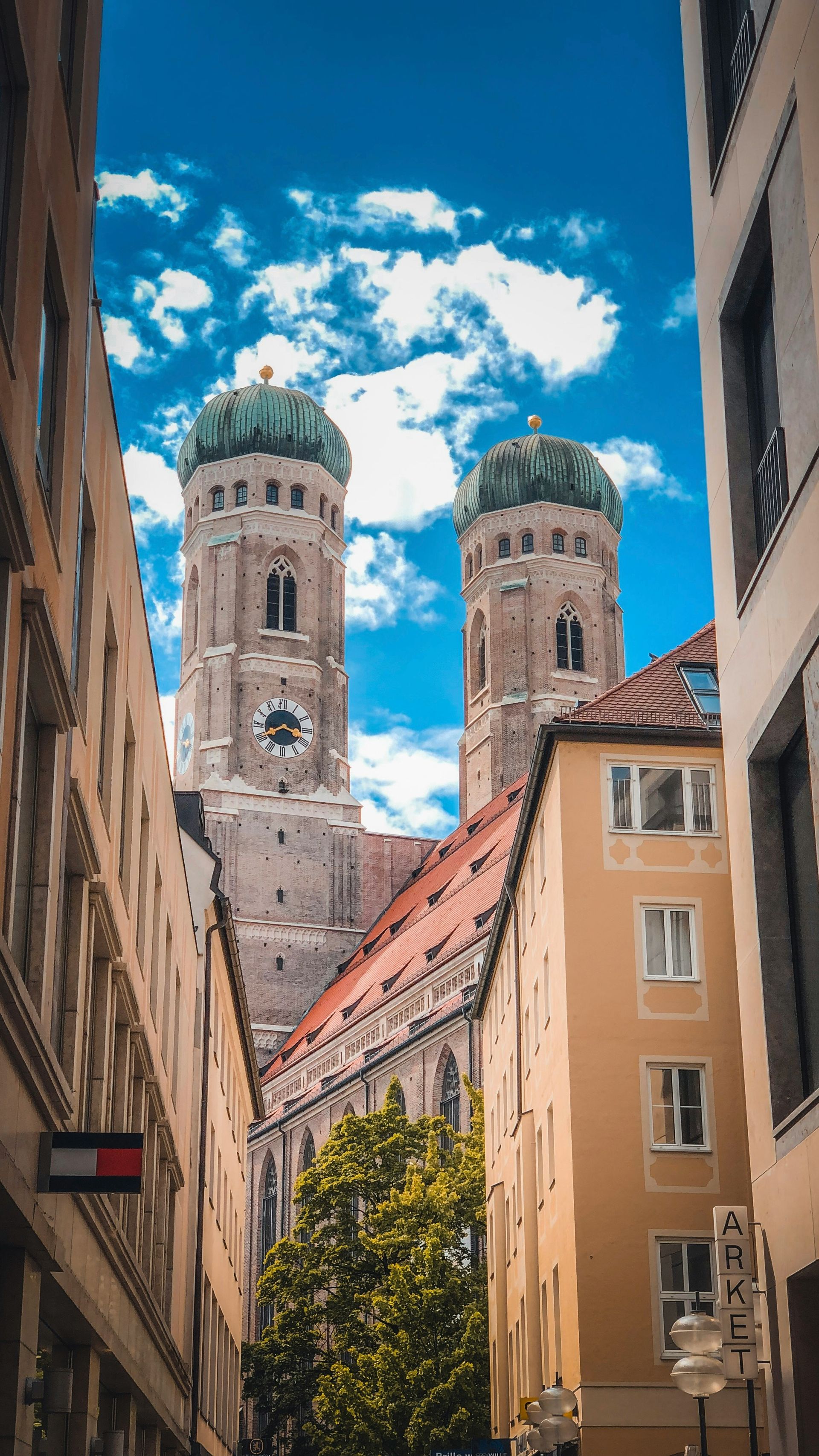 Towers of Frauenkirche church rise above buildings in Munich, Germany, under a blue sky with puffy clouds.