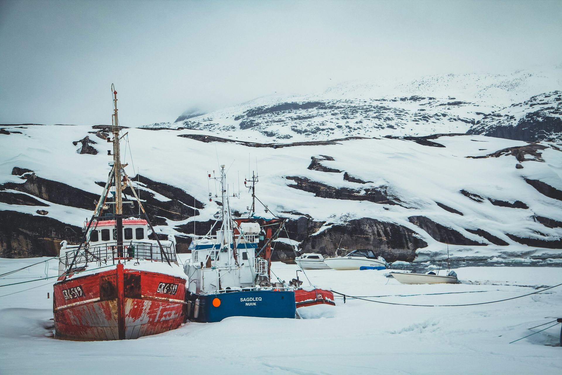 Red and blue boats docked on snowy shore; snow-covered hills in the background in Disko Bay, Greenland.