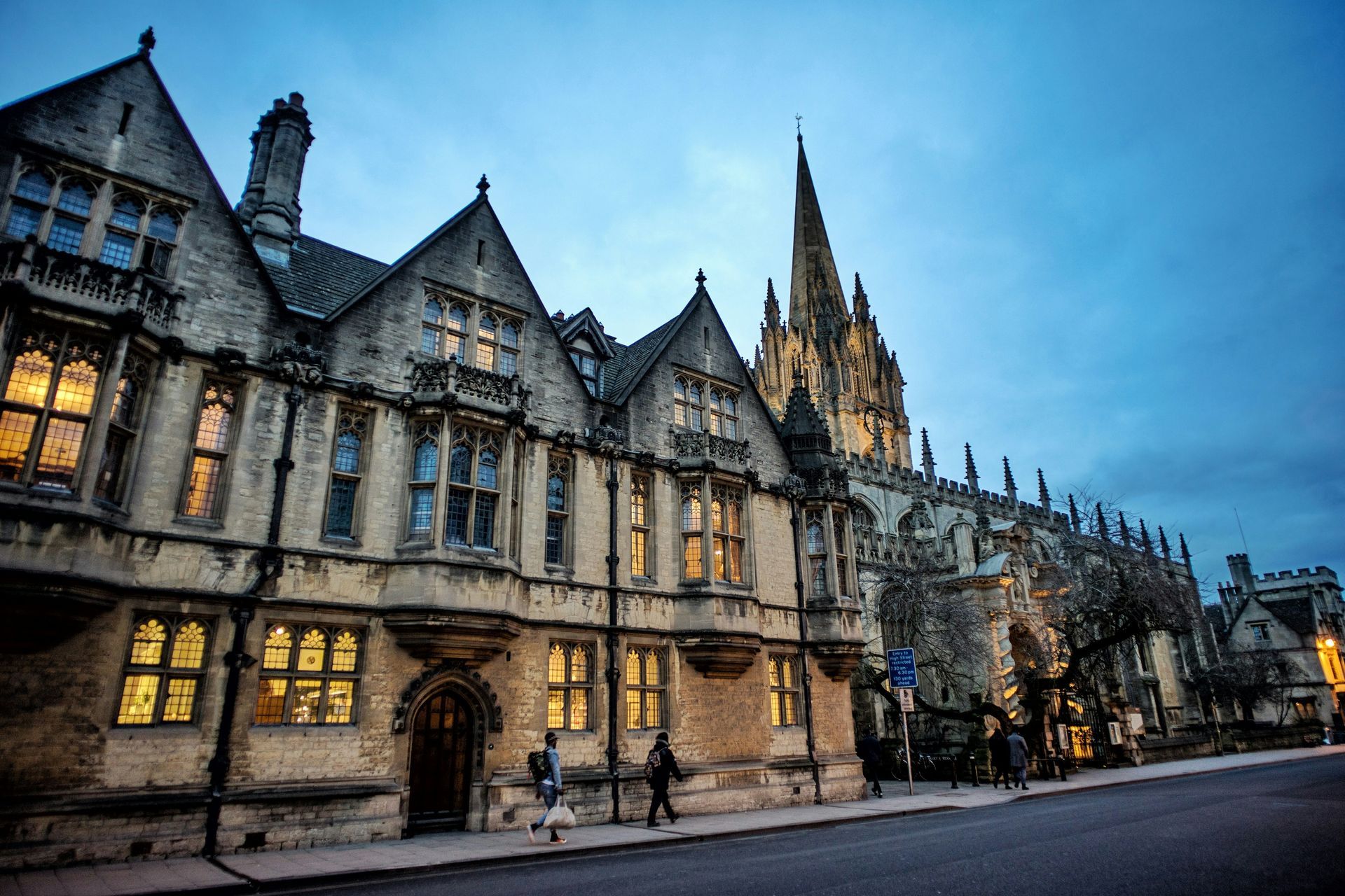 Stone buildings along a street at dusk; figures walk. Gothic spire rises in Oxford, England.