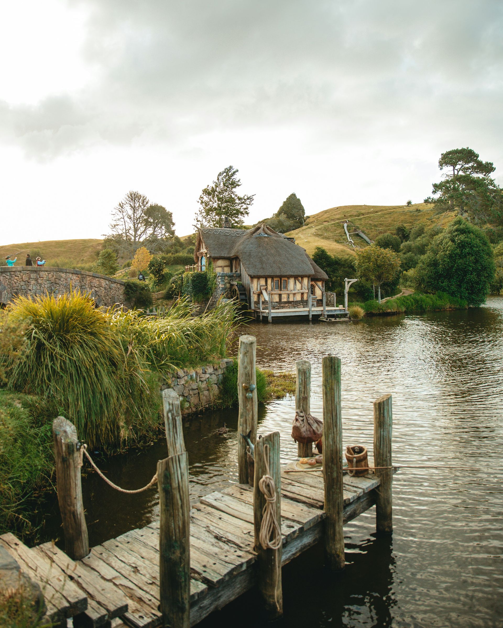 A wooden dock overlooking a pond with a house in the background at the Hobbiton Movie Set in Matamata, New Zealand.
