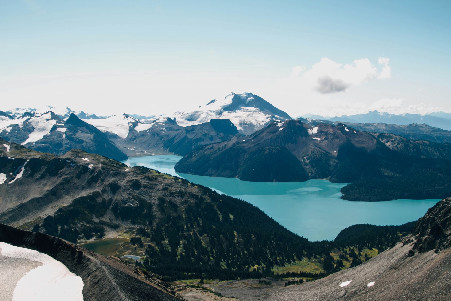 Panoramic view of a turquoise lake surrounded by snow-capped mountains and dark green forests in Garibaldi Park, Canada.