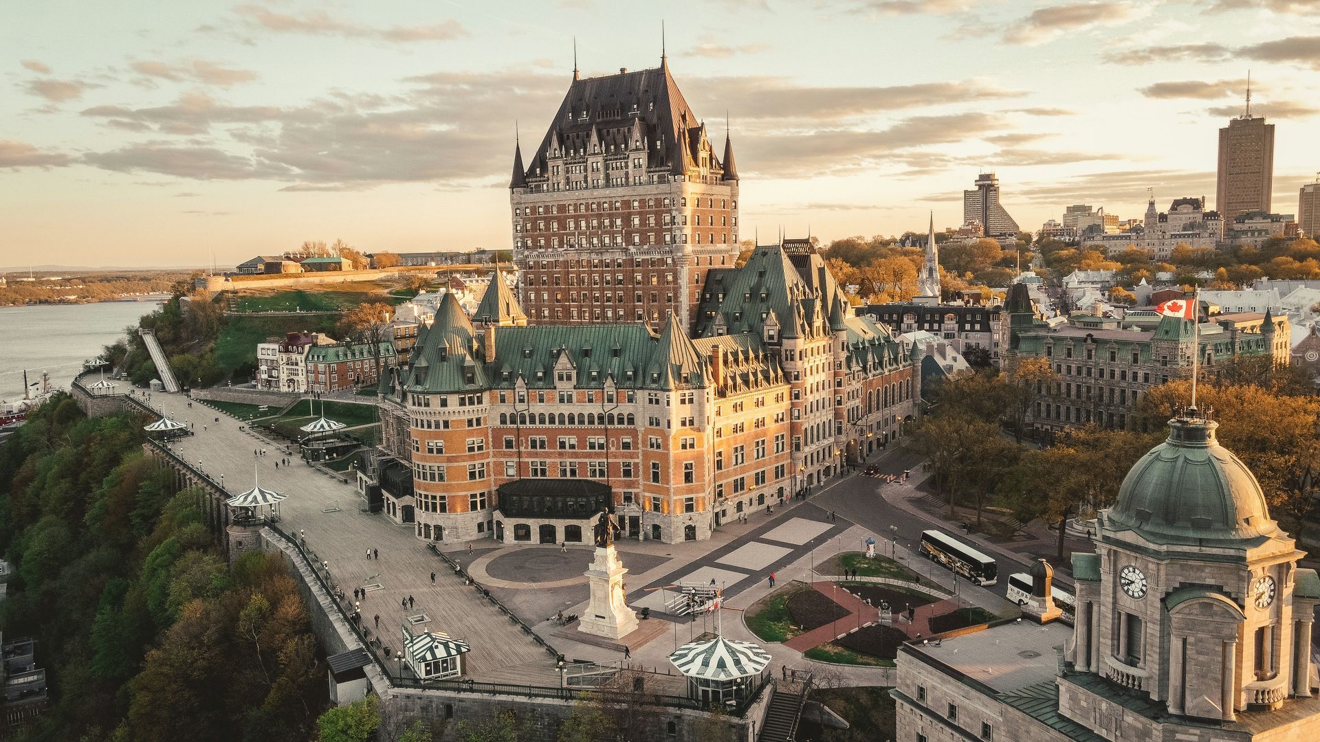 Fairmont Le Château Frontenac, a large castle-like hotel, overlooks Quebec City. A tree-lined path, monument, and river are in the foreground.