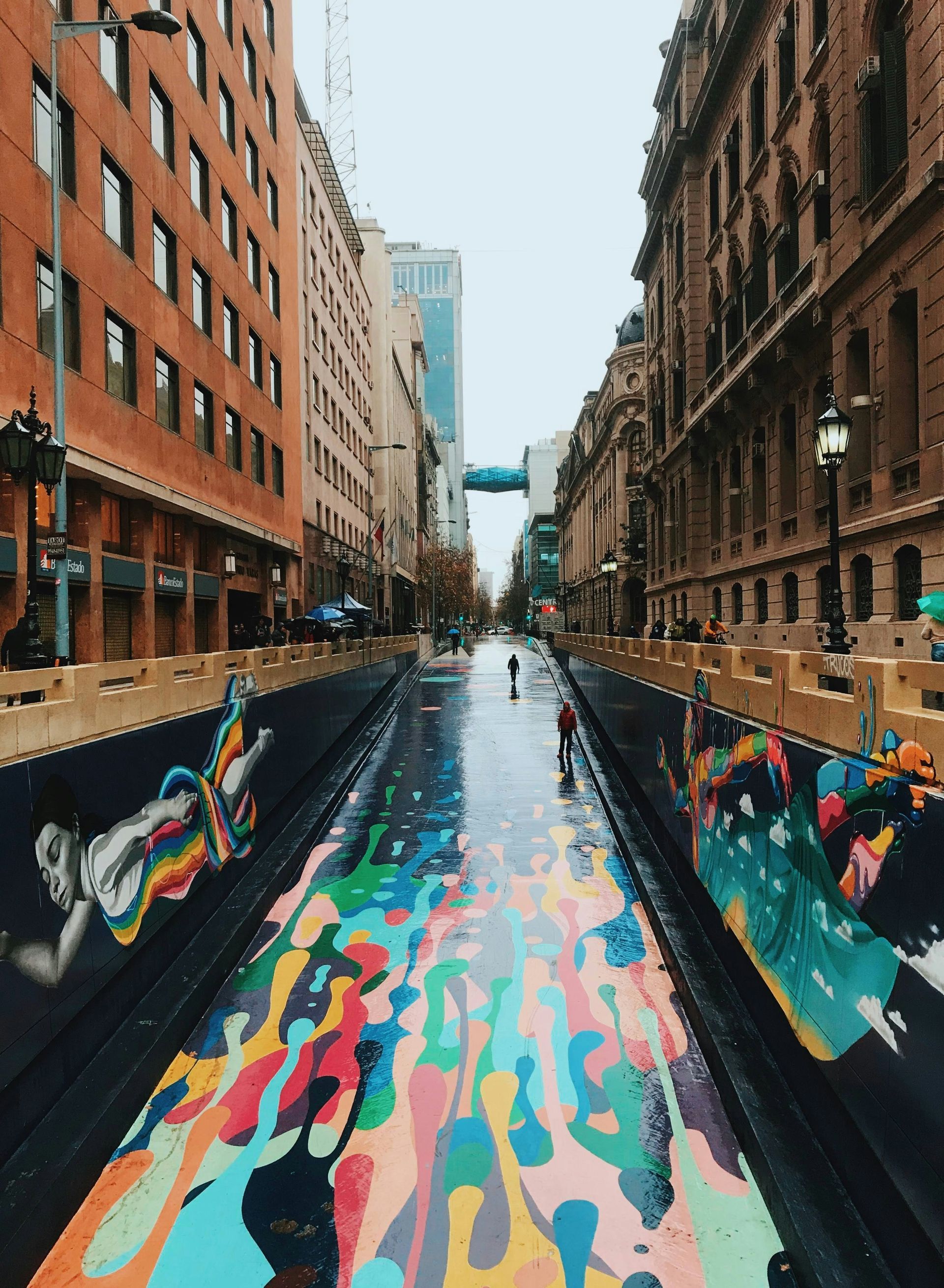 Street art mural with colorful paint drips on a wet road, flanked by brick buildings in Santiago, Chile.
