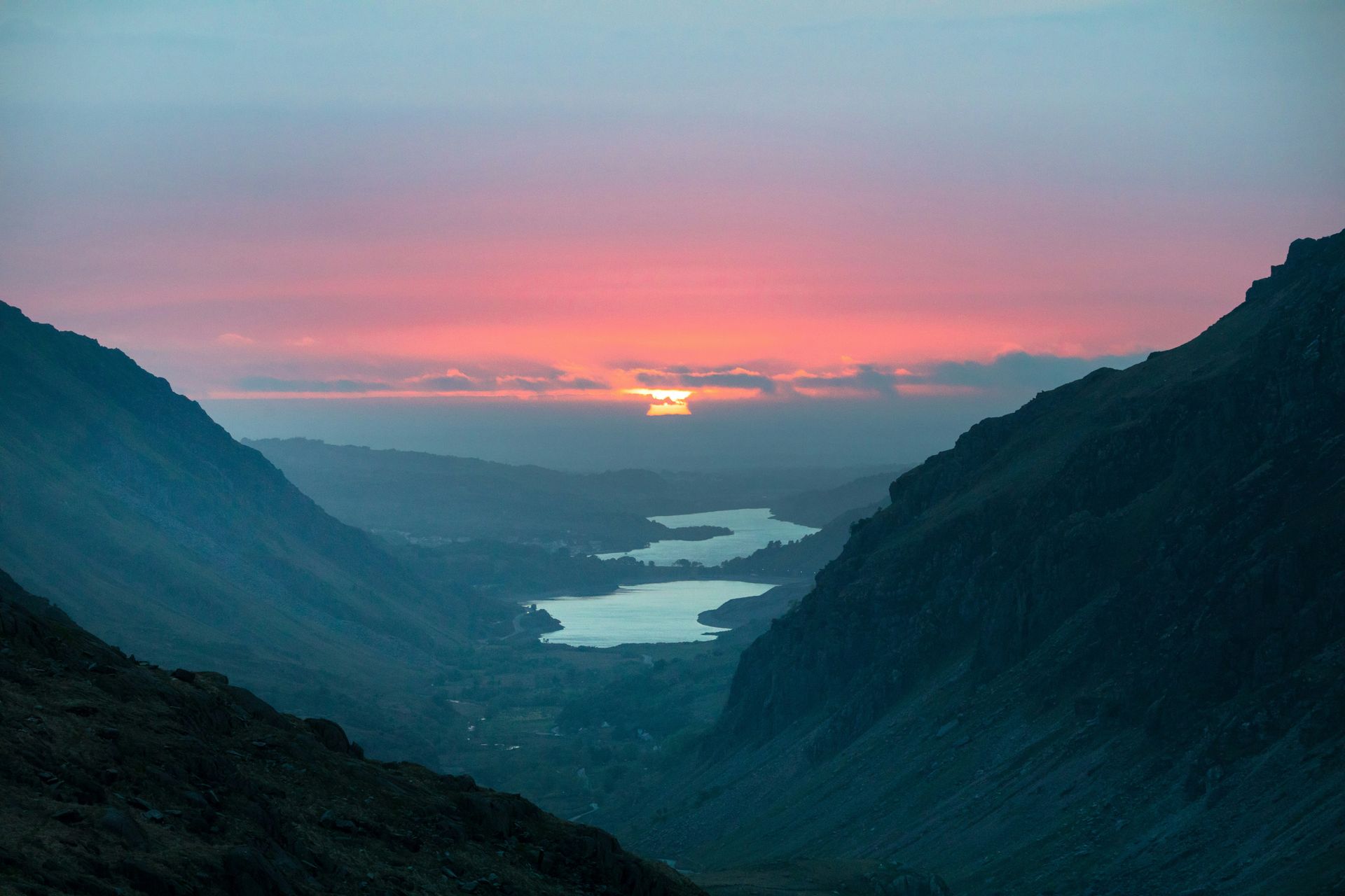 Sunset over a valley with two lakes, framed by dark mountain peaks, pink and orange sky in Snowdonia, Wales.