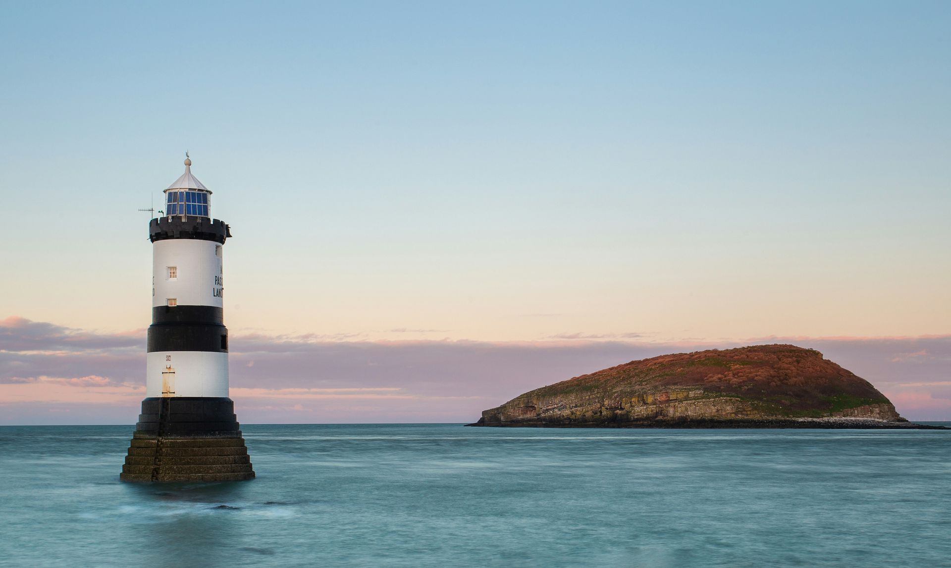 Lighthouse with black and white stripes next to a small, green island in calm water under a dusky sky in Anglesey, Wales