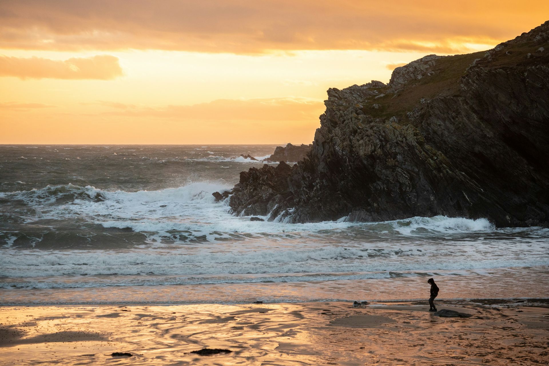 Person on beach watches waves crash against rocky cliff at sunset in Anglesey, Wales
