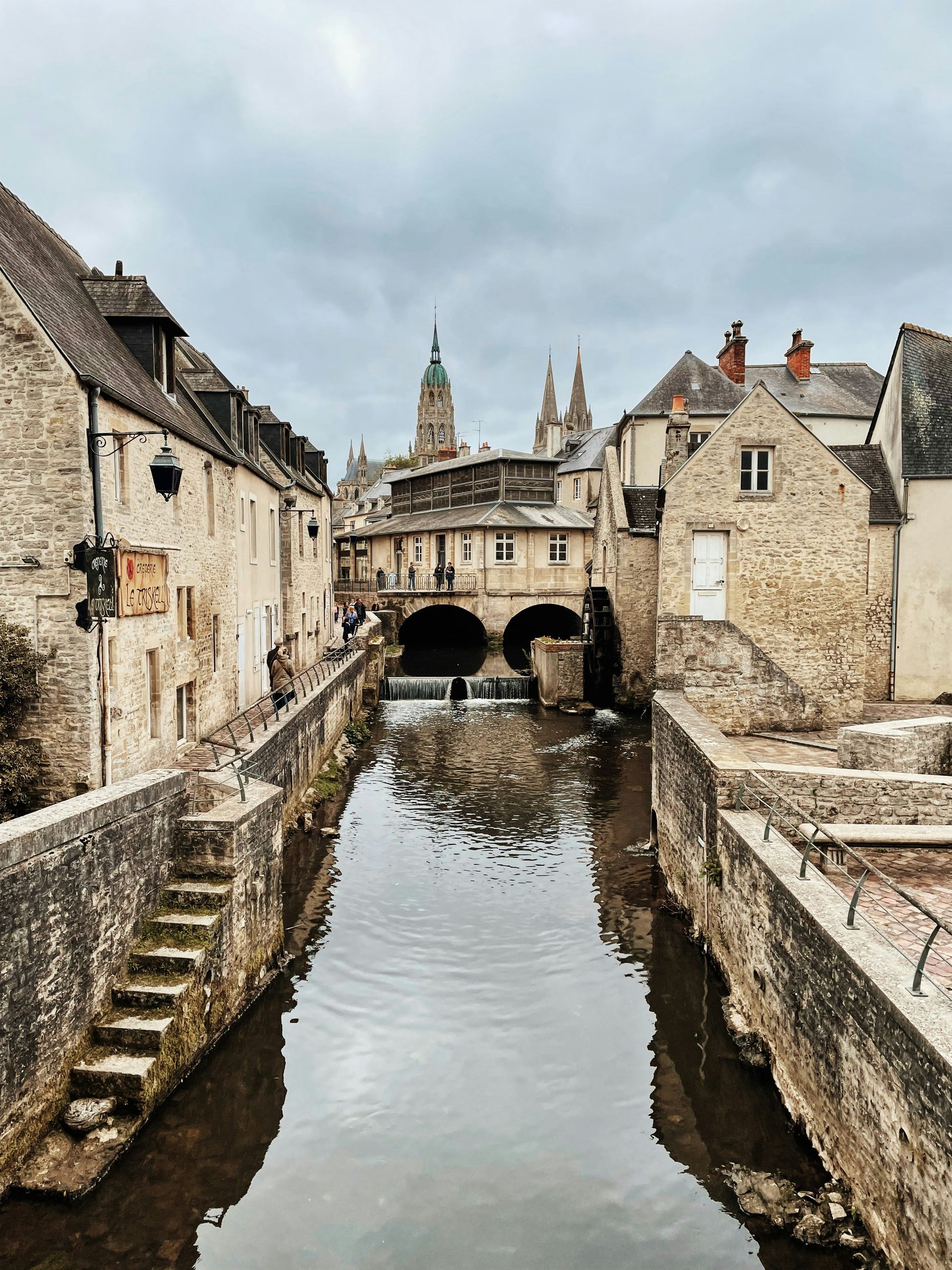 A river runs through a small town surrounded by stone buildings in Normandy, France.