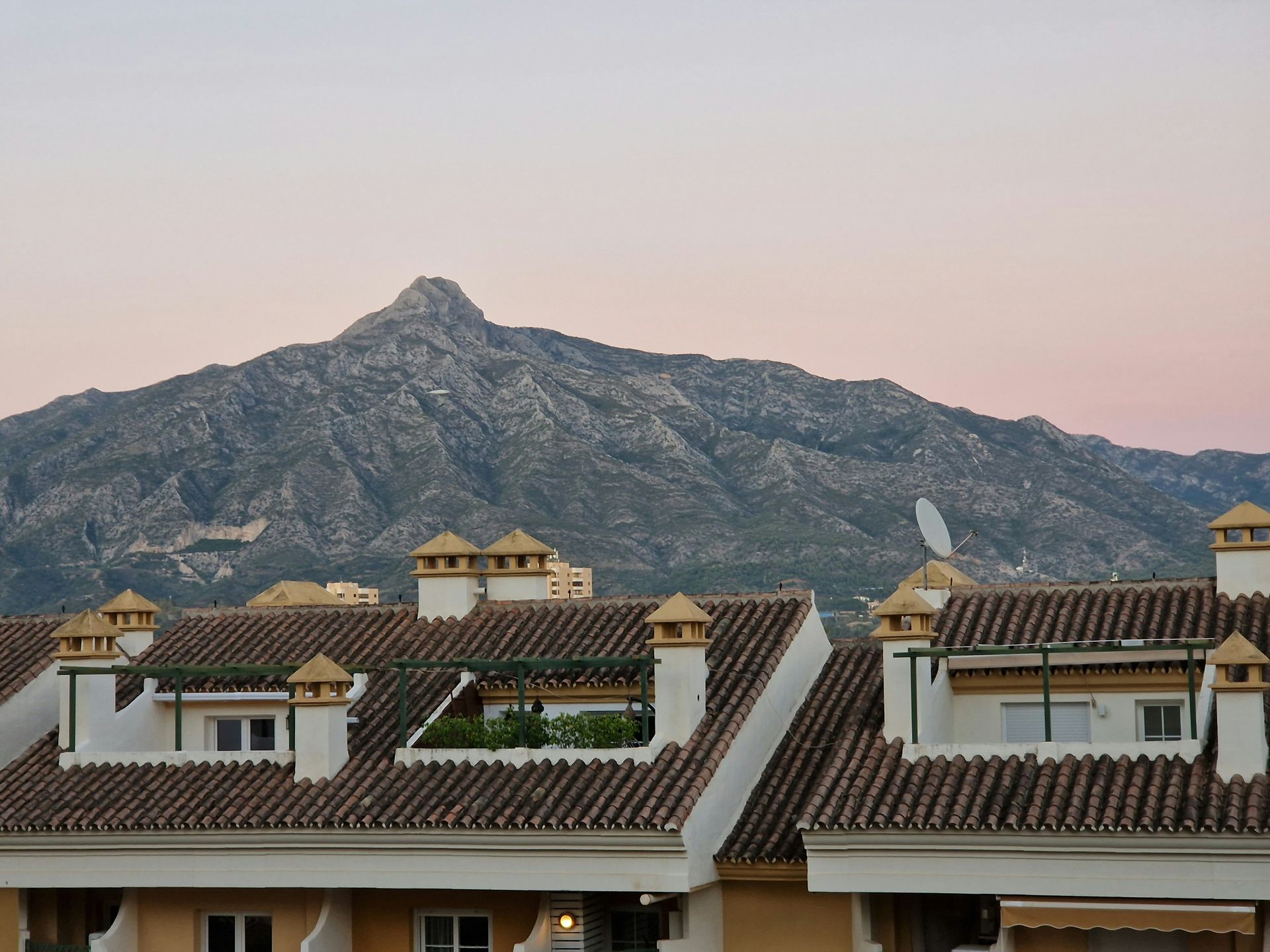 Rooftops with chimneys in front of a large, rocky mountain under a pink and purple sky in Marbella, Spain.