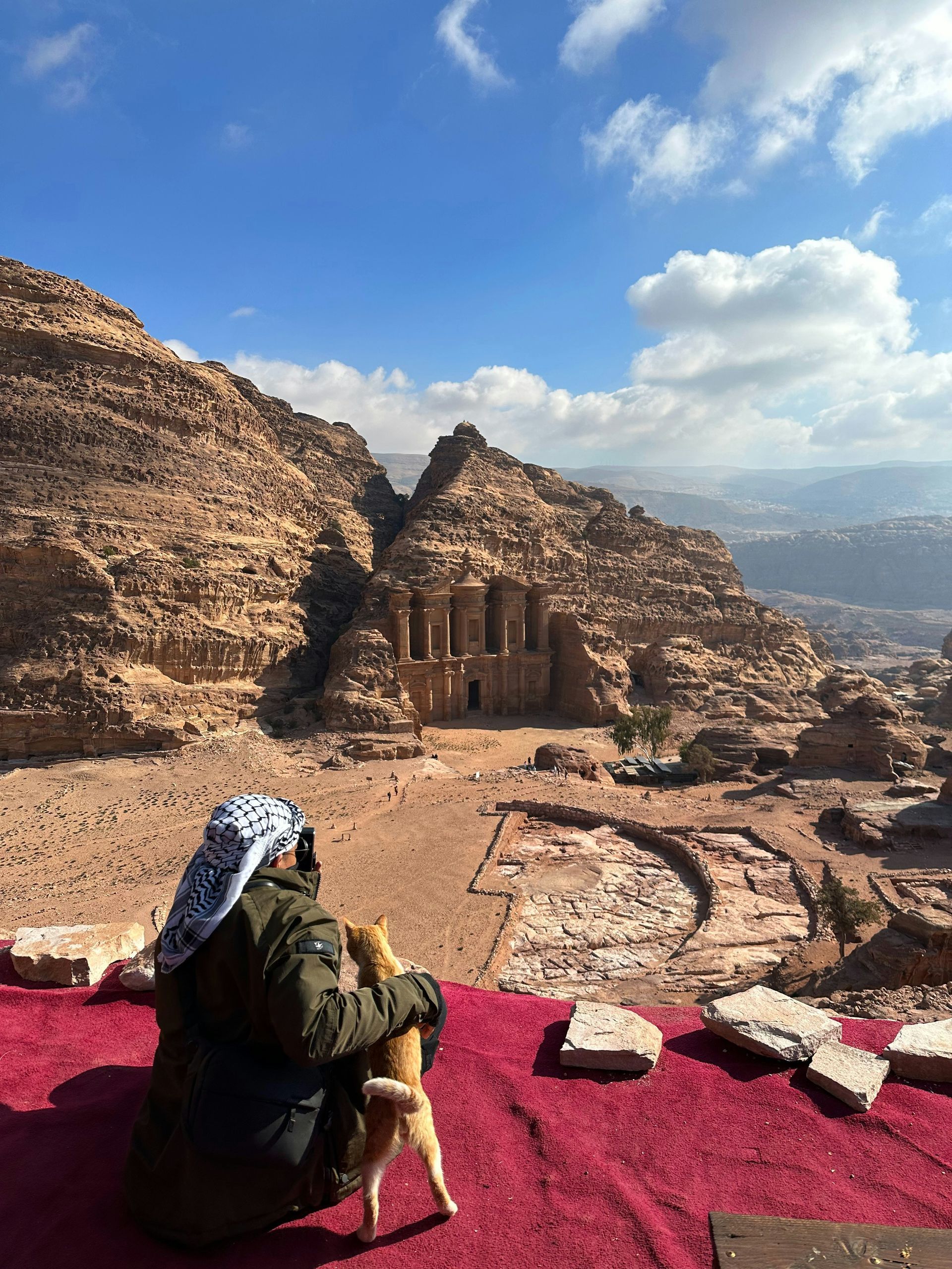 Person in traditional head covering sits with a dog, gazing at Petra's Monastery, carved into sandstone cliffs, under a blue sky.