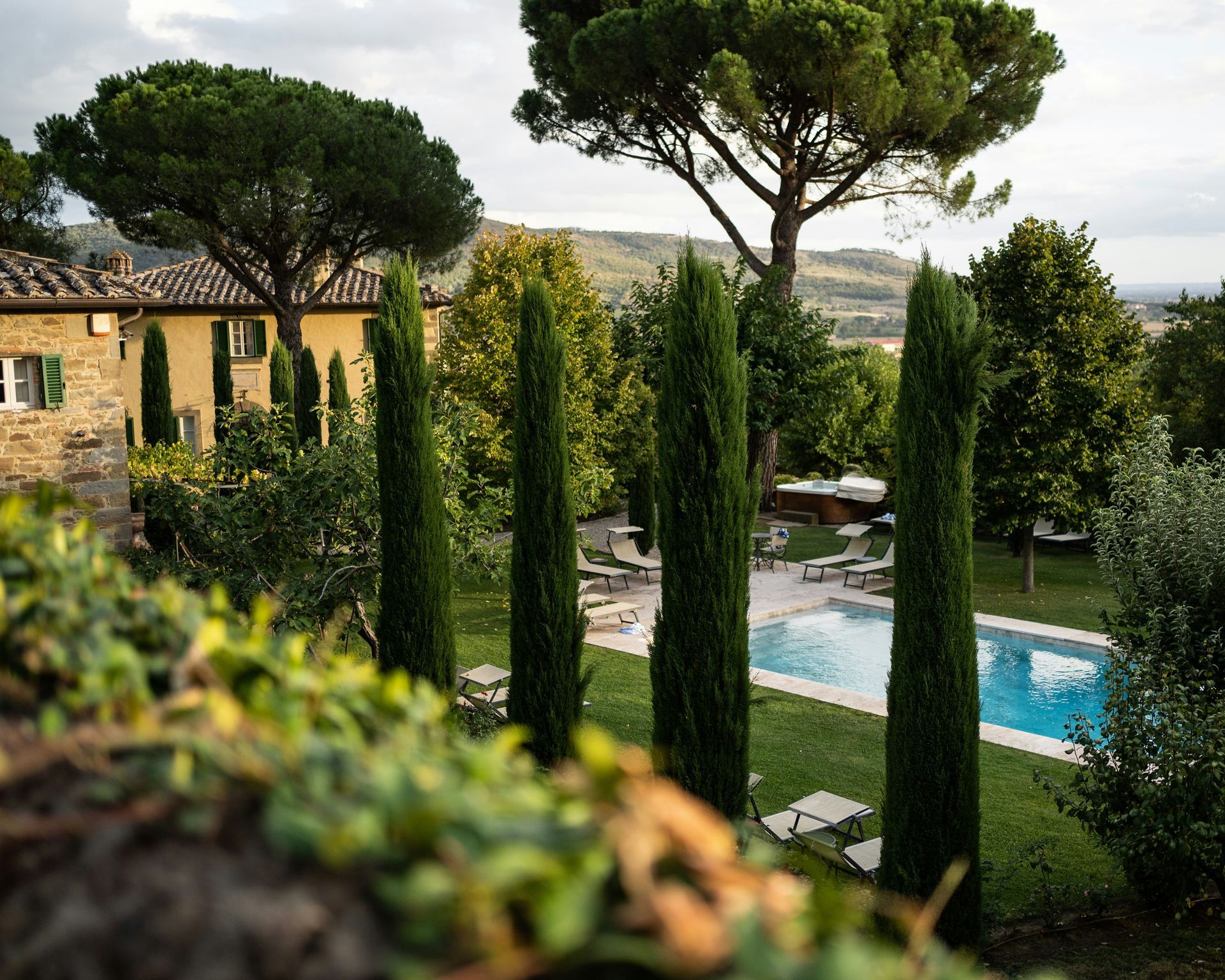 A swimming pool is surrounded by trees and a house in the background in Asciano, Tuscany.