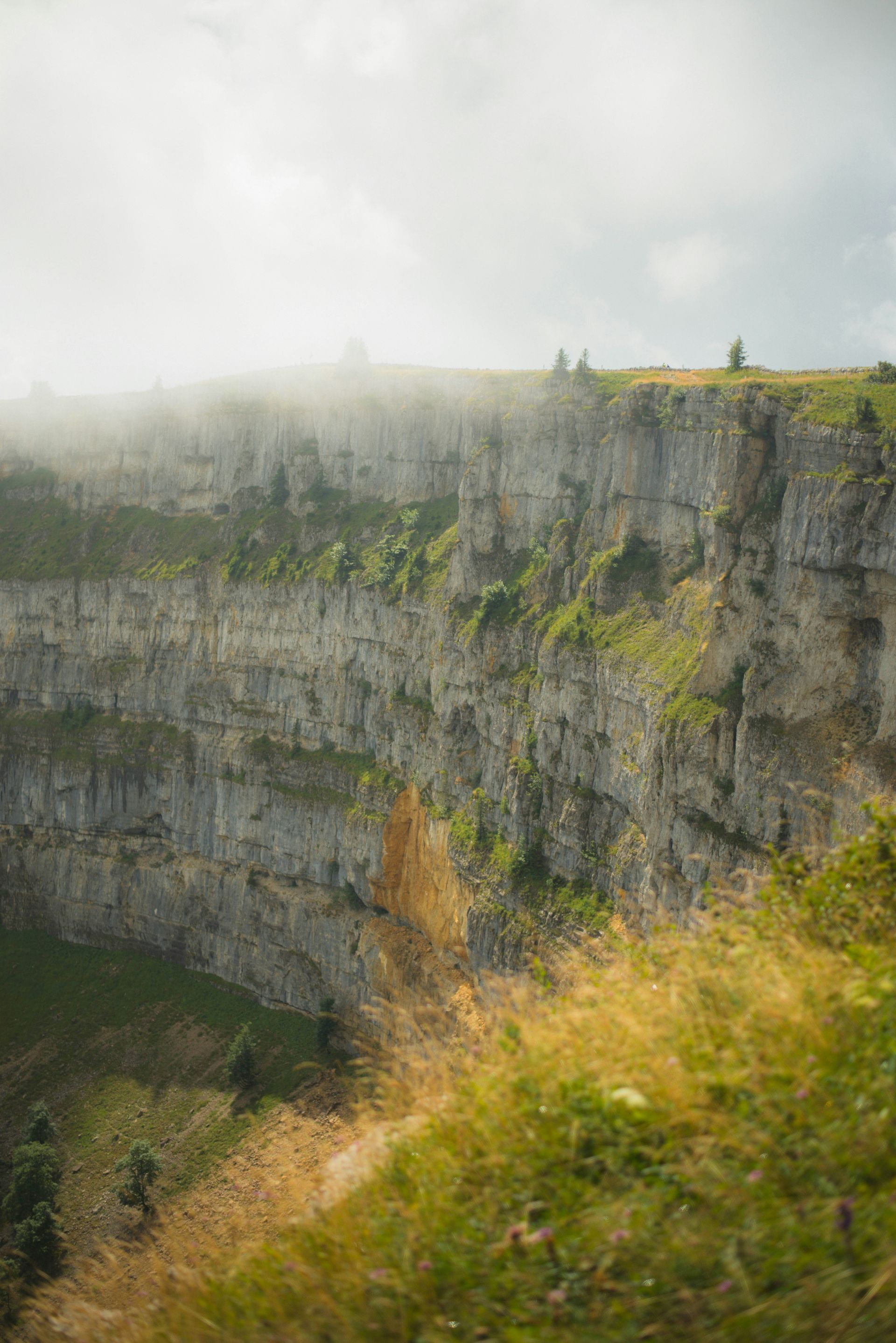 Rocky cliff face of Creux du Van with grass and some trees. Fog hangs at the top in Switzerland. 