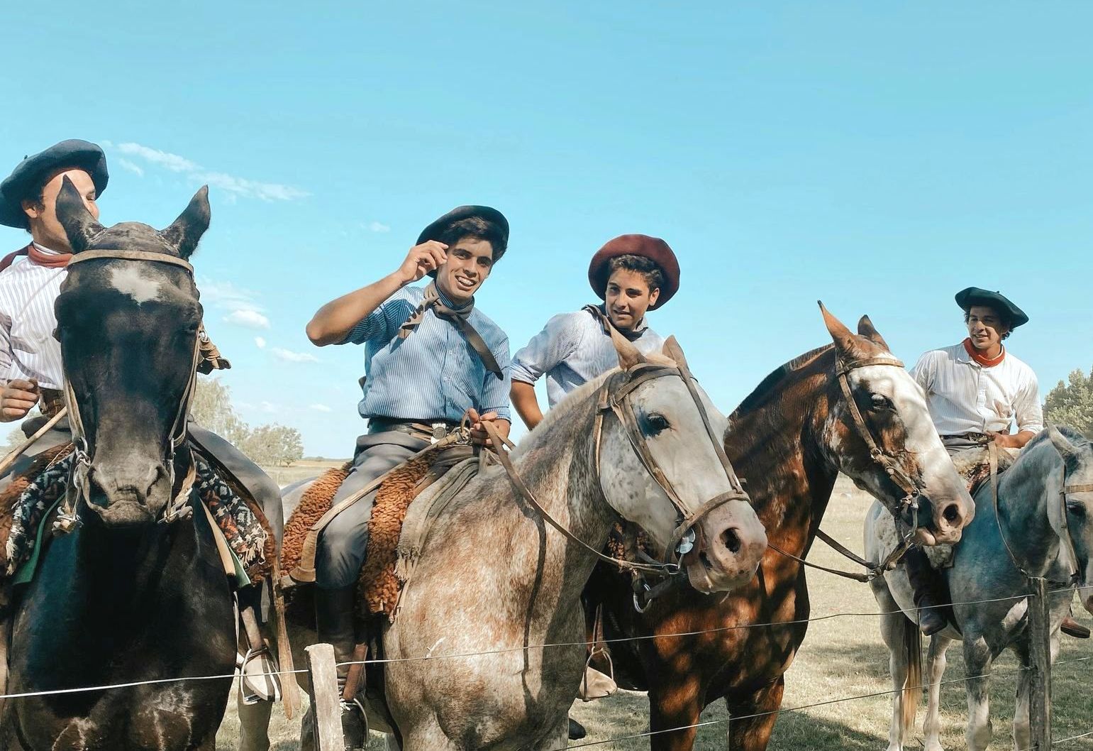 Five gauchos on horseback, wearing hats and traditional clothing, smiling in a field under a blue sky in Argentina.