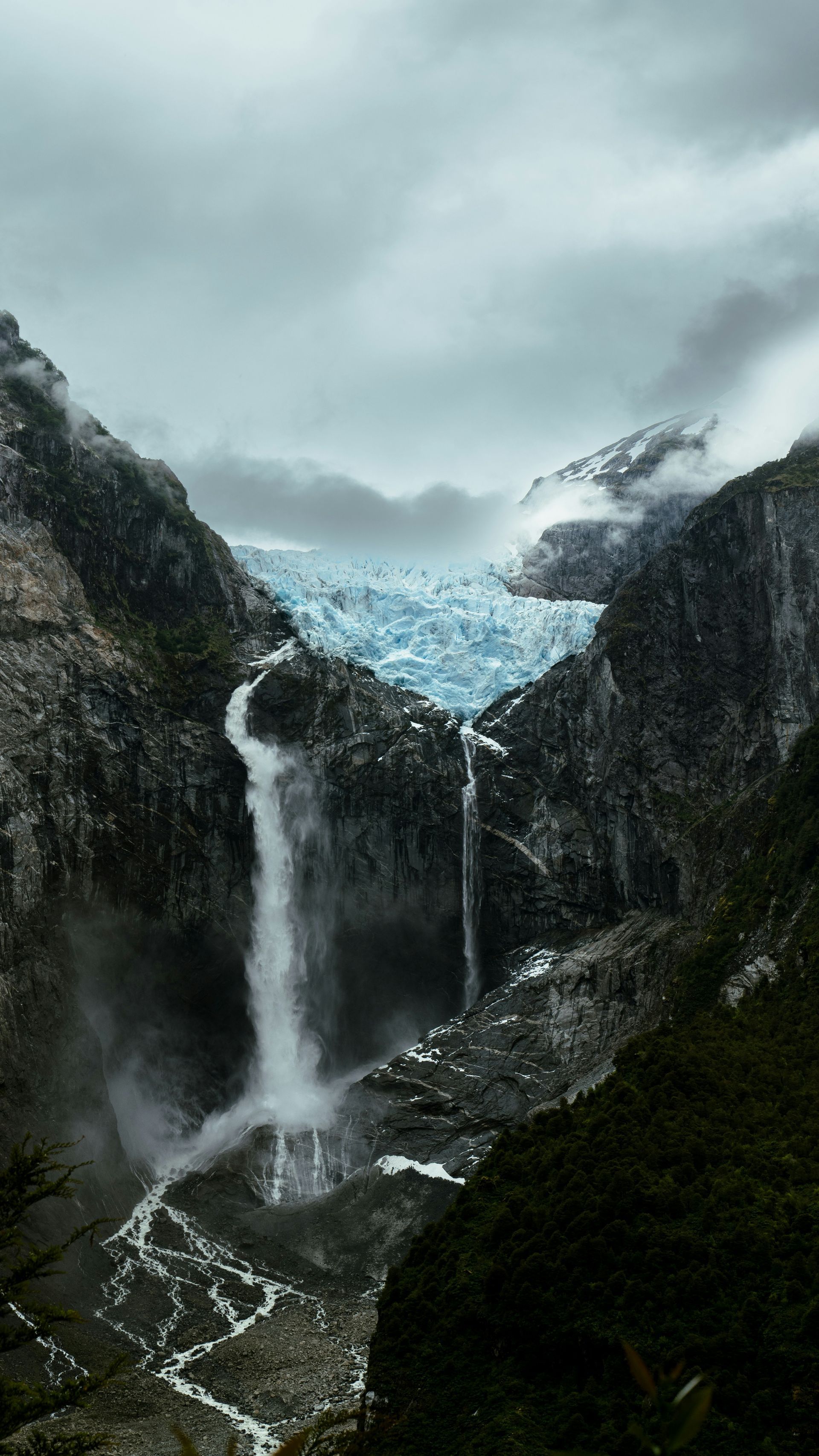 Waterfall cascading from a glacier in a rocky mountain setting under a cloudy sky at Parque Nacional Queulat, Chile.